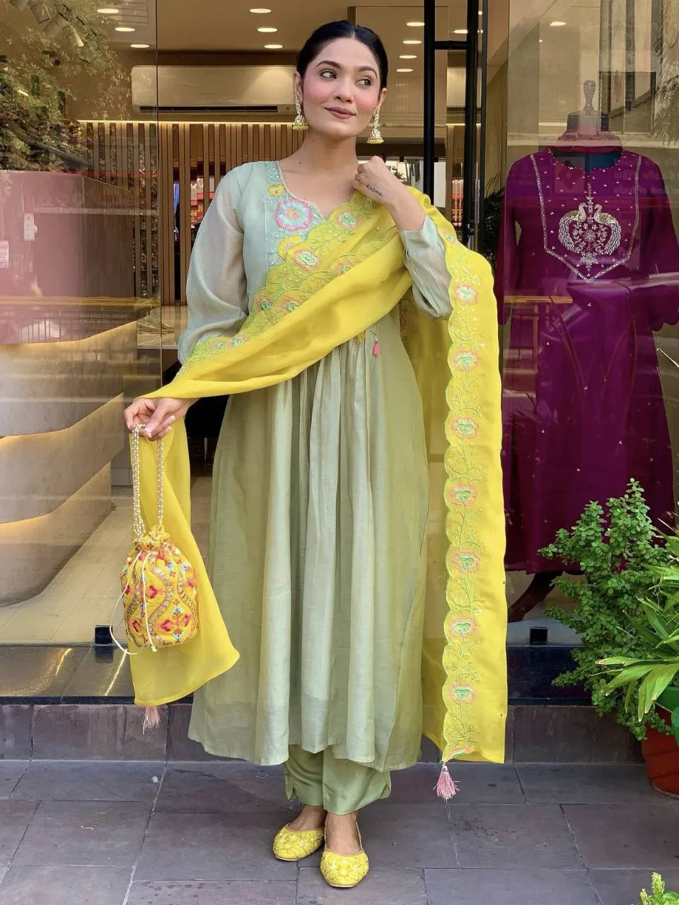 Woman in a light green traditional outfit with a yellow dupatta standing in front of a store.