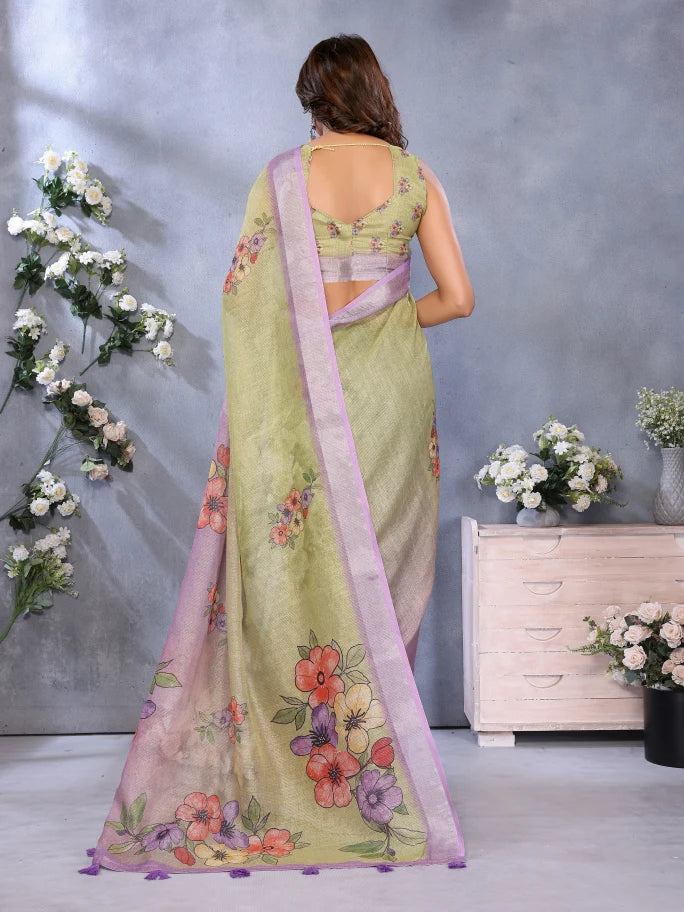 Woman wearing a light green saree with floral patterns in a room with decorative flowers and furniture.