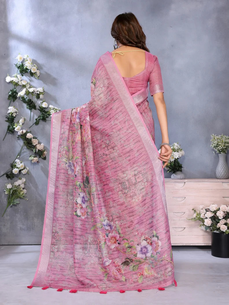 Woman wearing a pink saree with floral patterns in a decorative setting with flowers and a wooden cabinet.