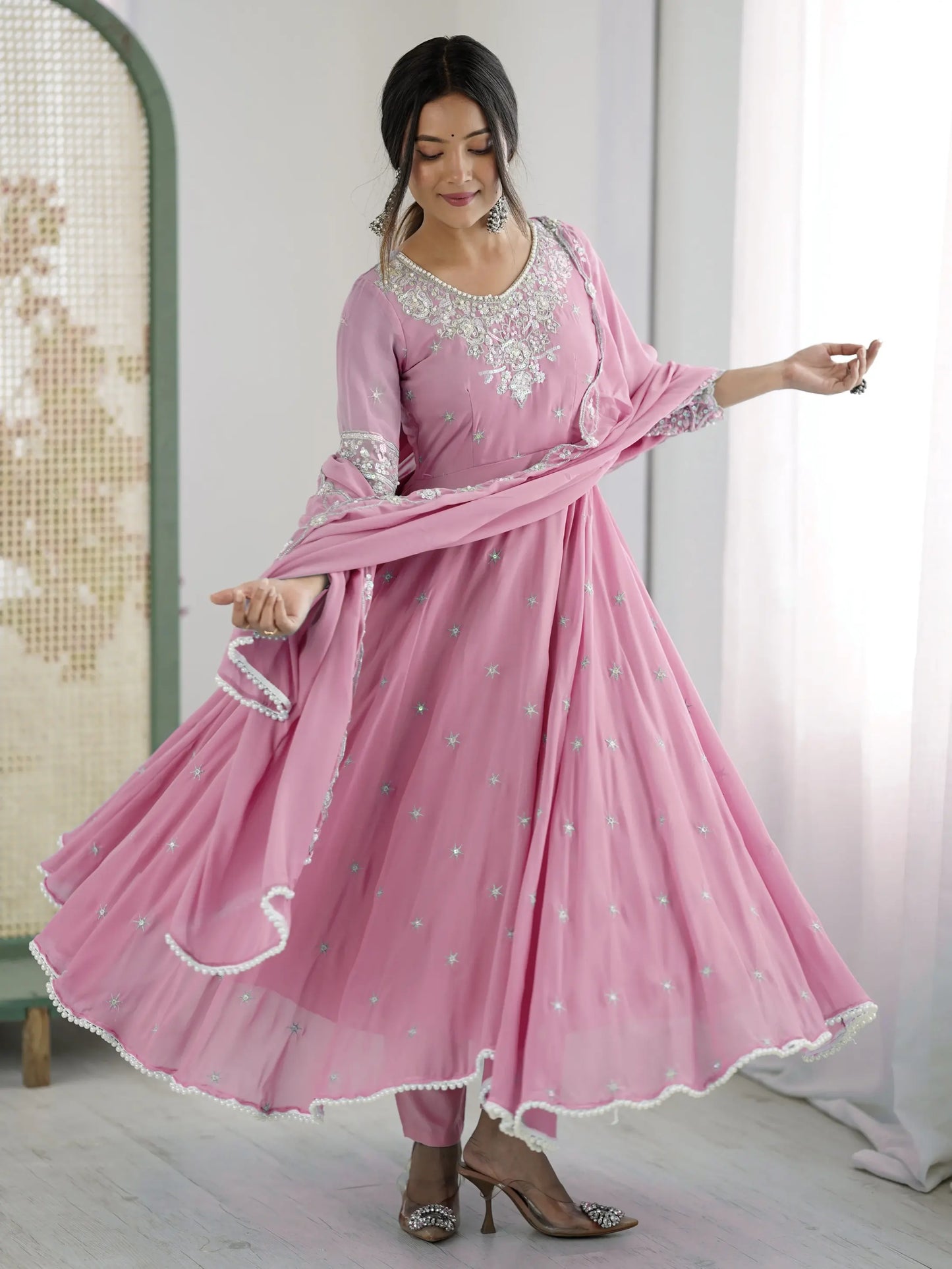 Woman wearing a pink traditional outfit with white embroidery in a minimal indoor setting.