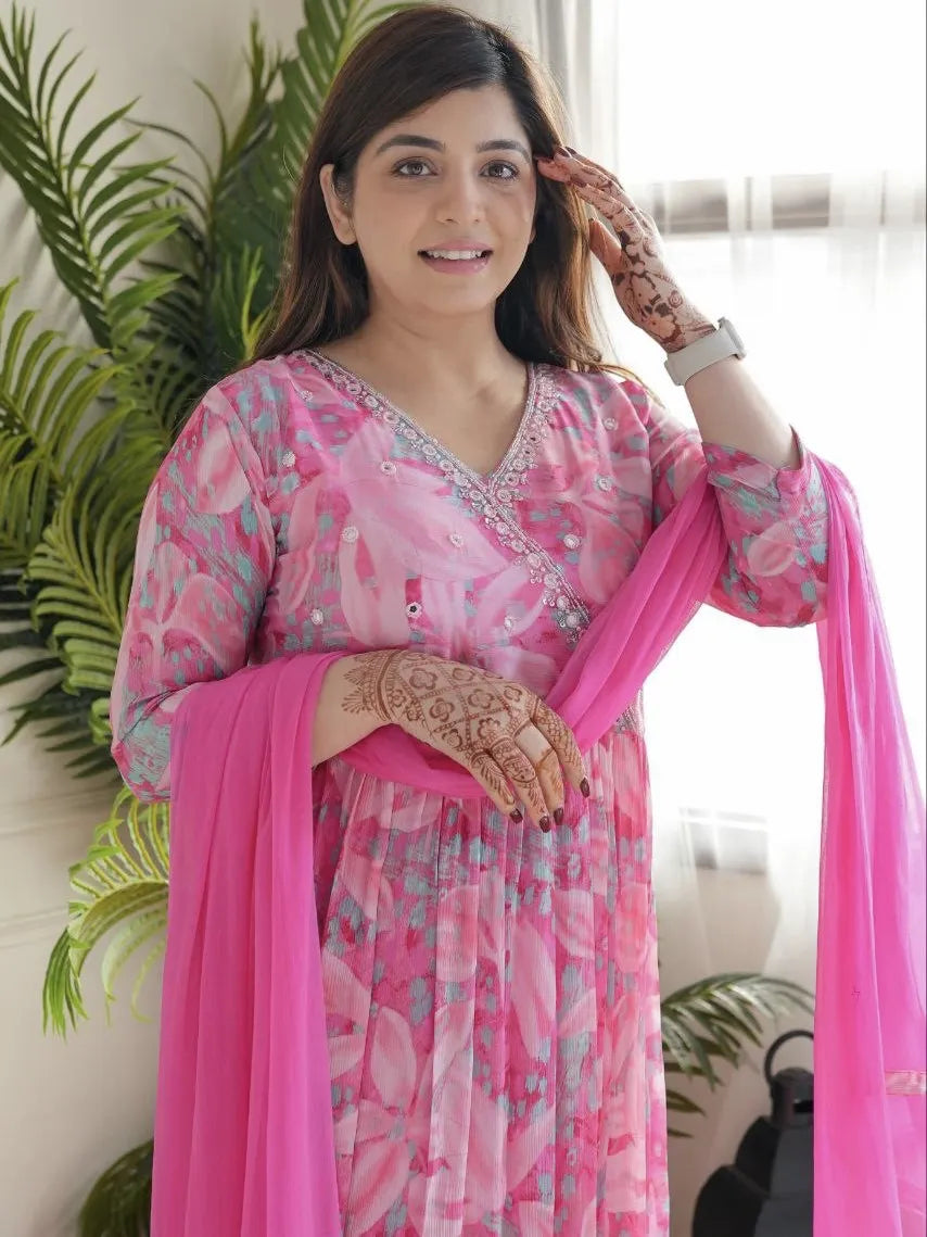 Woman in a pink traditional outfit with floral patterns, standing indoors with plants in the background.