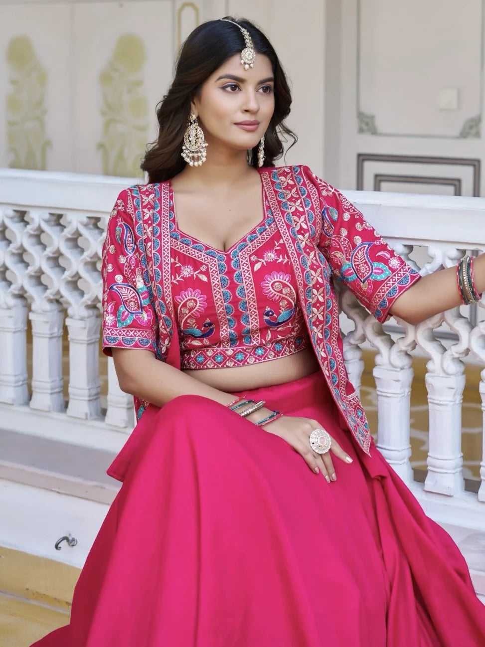 Woman in a pink traditional outfit with intricate patterns, sitting on a decorative balcony.