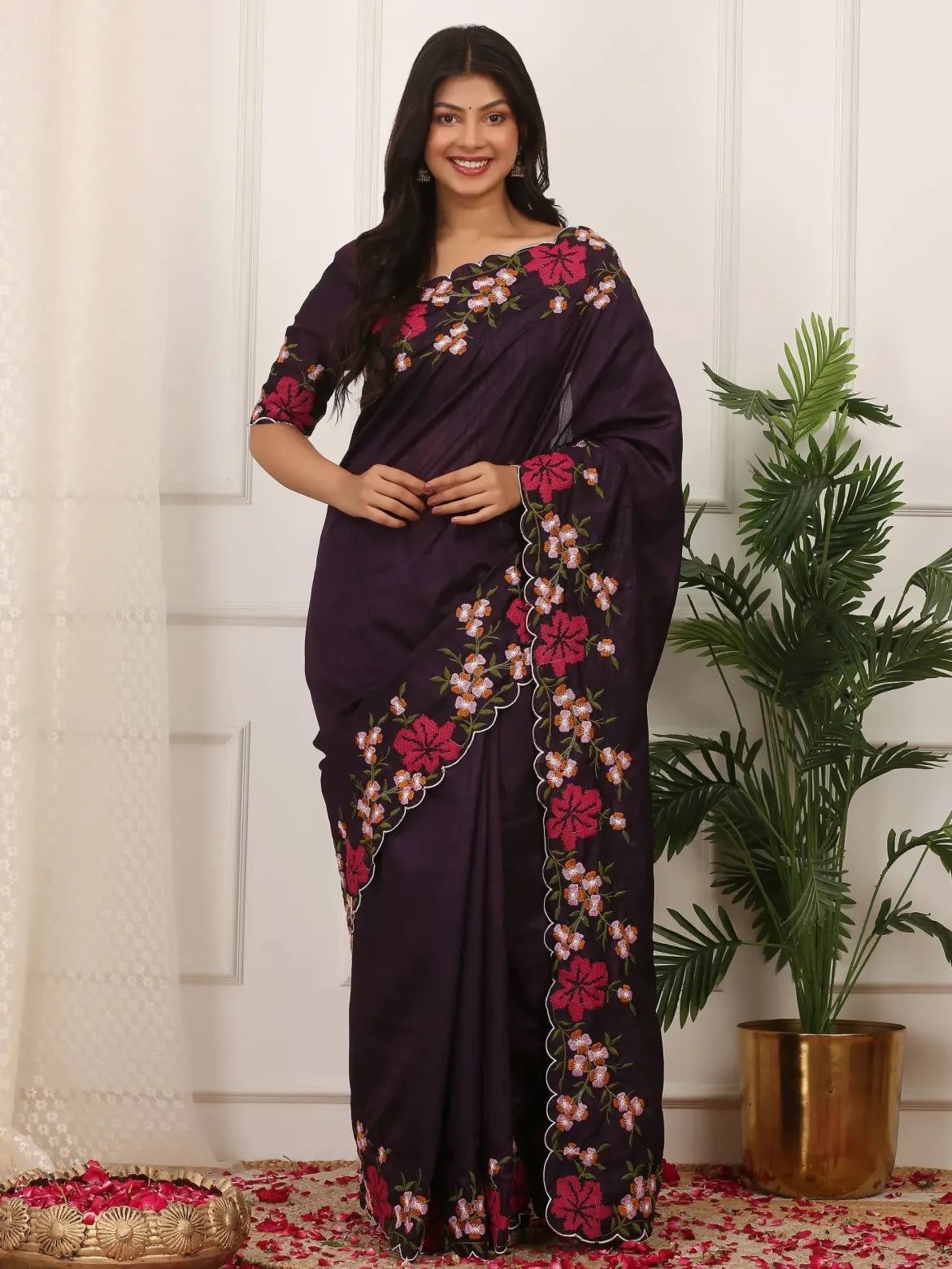 Woman wearing a dark purple saree with floral embroidery in a decorated indoor setting.