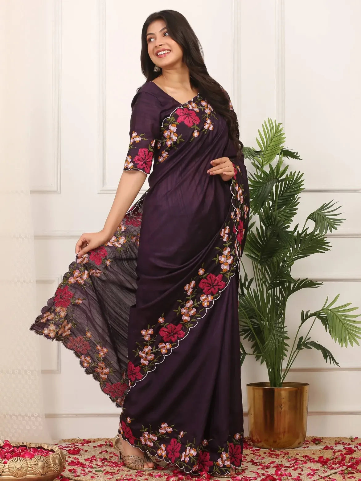 Woman wearing a dark purple saree with floral patterns, standing indoors next to a plant.