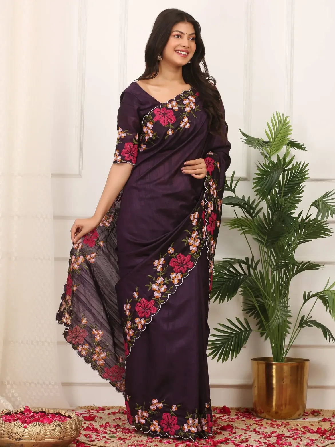 Woman wearing a dark purple saree with floral patterns in a room with a plant and decorative items.