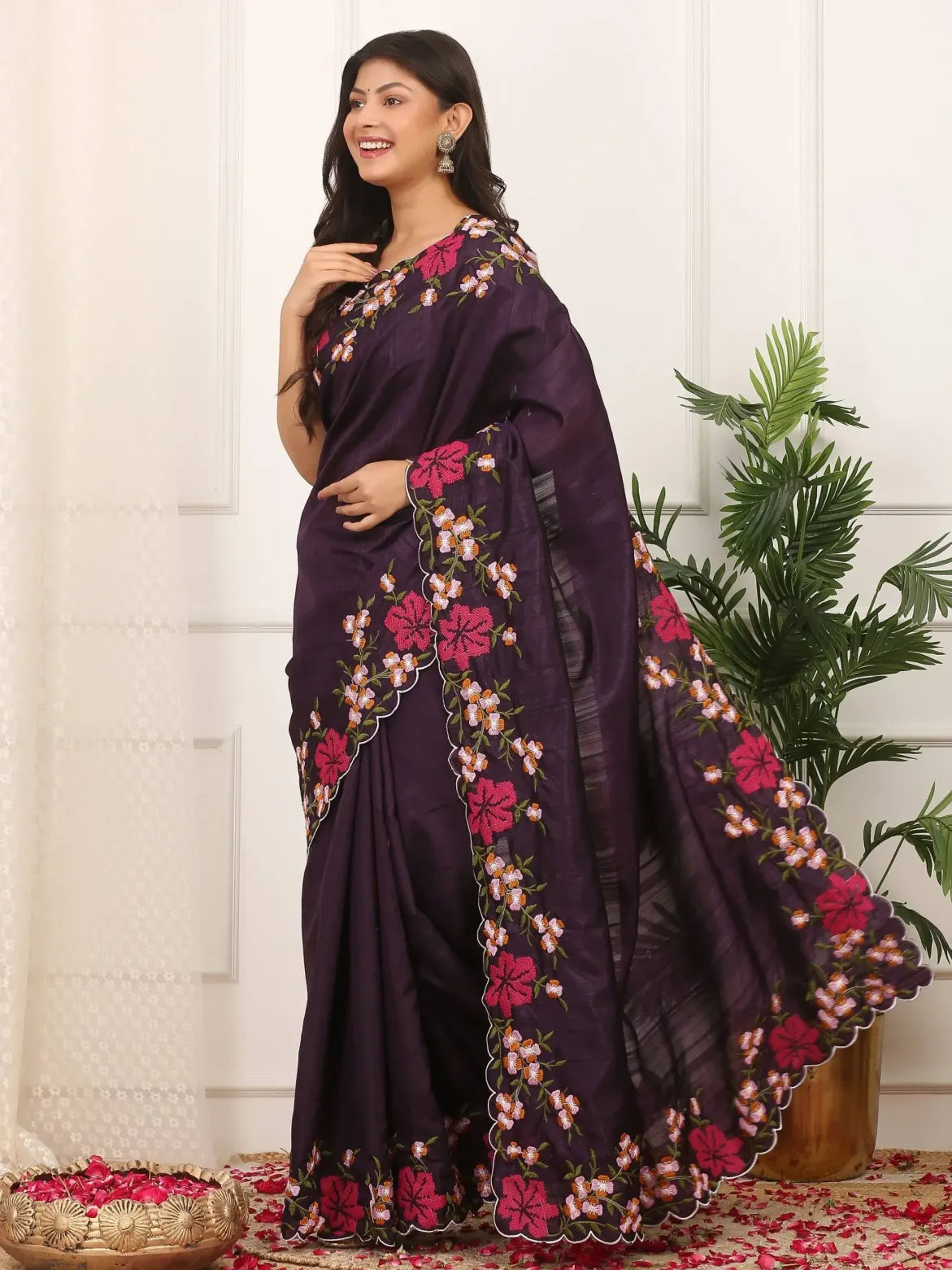 Woman wearing a dark purple saree with floral embroidery in a room with white walls and a plant.