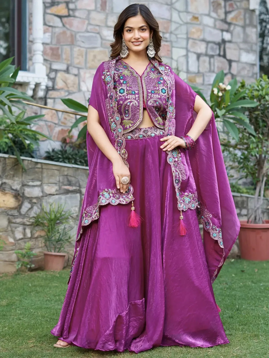 Woman in a purple traditional outfit with embroidery standing outdoors against a stone wall.