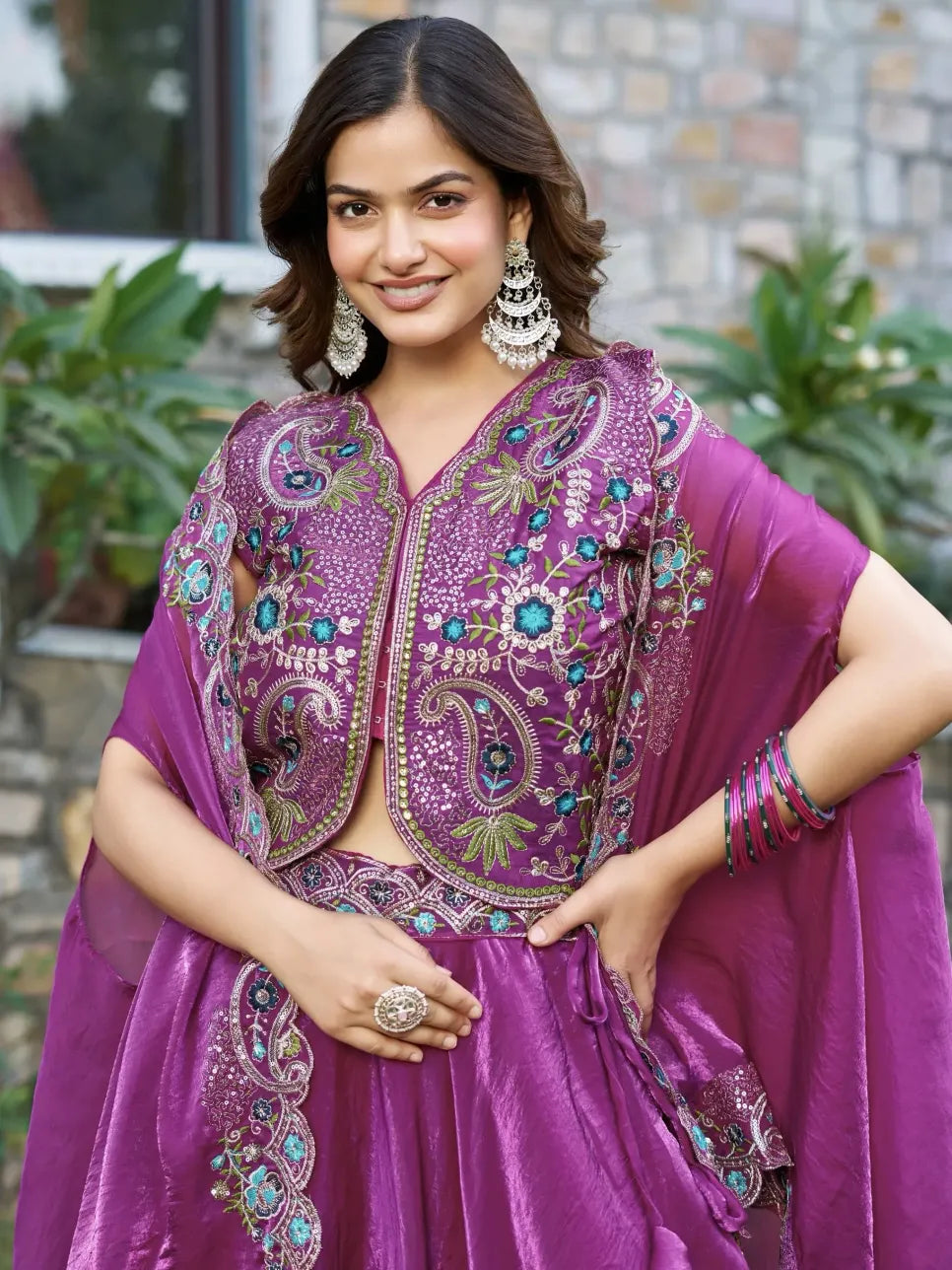 Woman wearing a purple embroidered traditional outfit with a stone wall and plants in the background