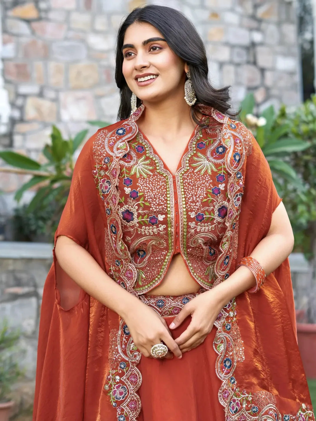 Woman wearing a traditional embroidered outfit with a stone wall and plants in the background