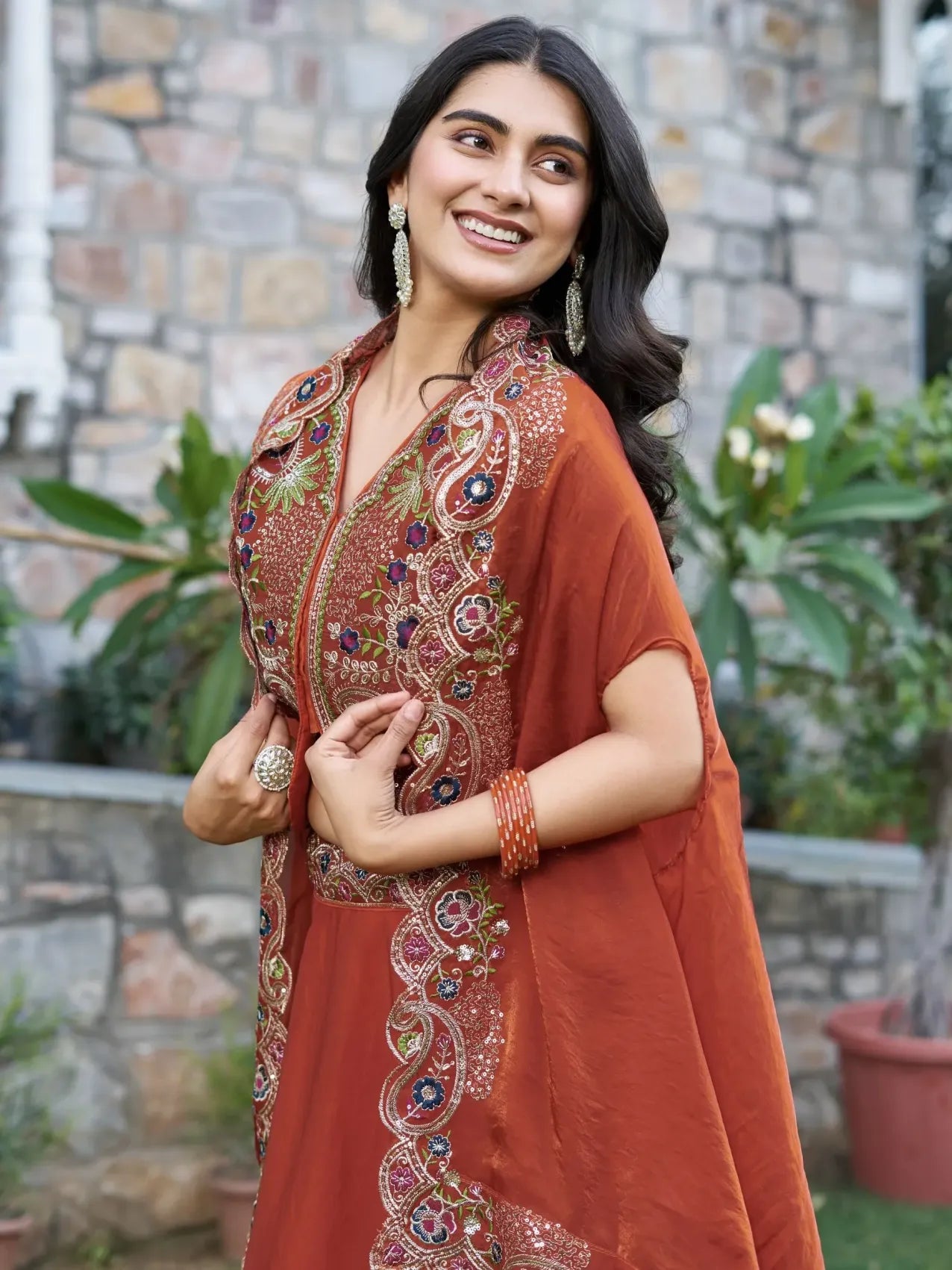 Woman wearing a traditional embroidered outfit with a stone wall and plants in the background