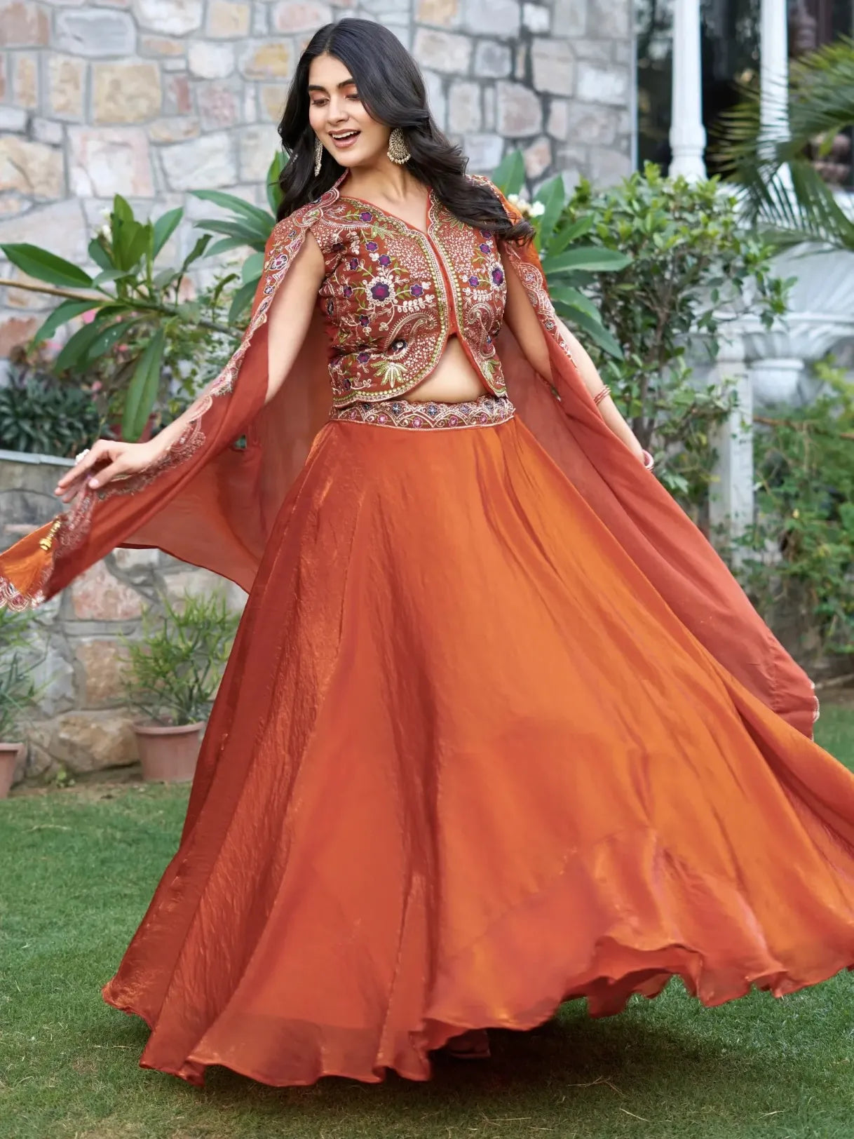 Woman in an orange traditional outfit with a stone wall and plants in the background