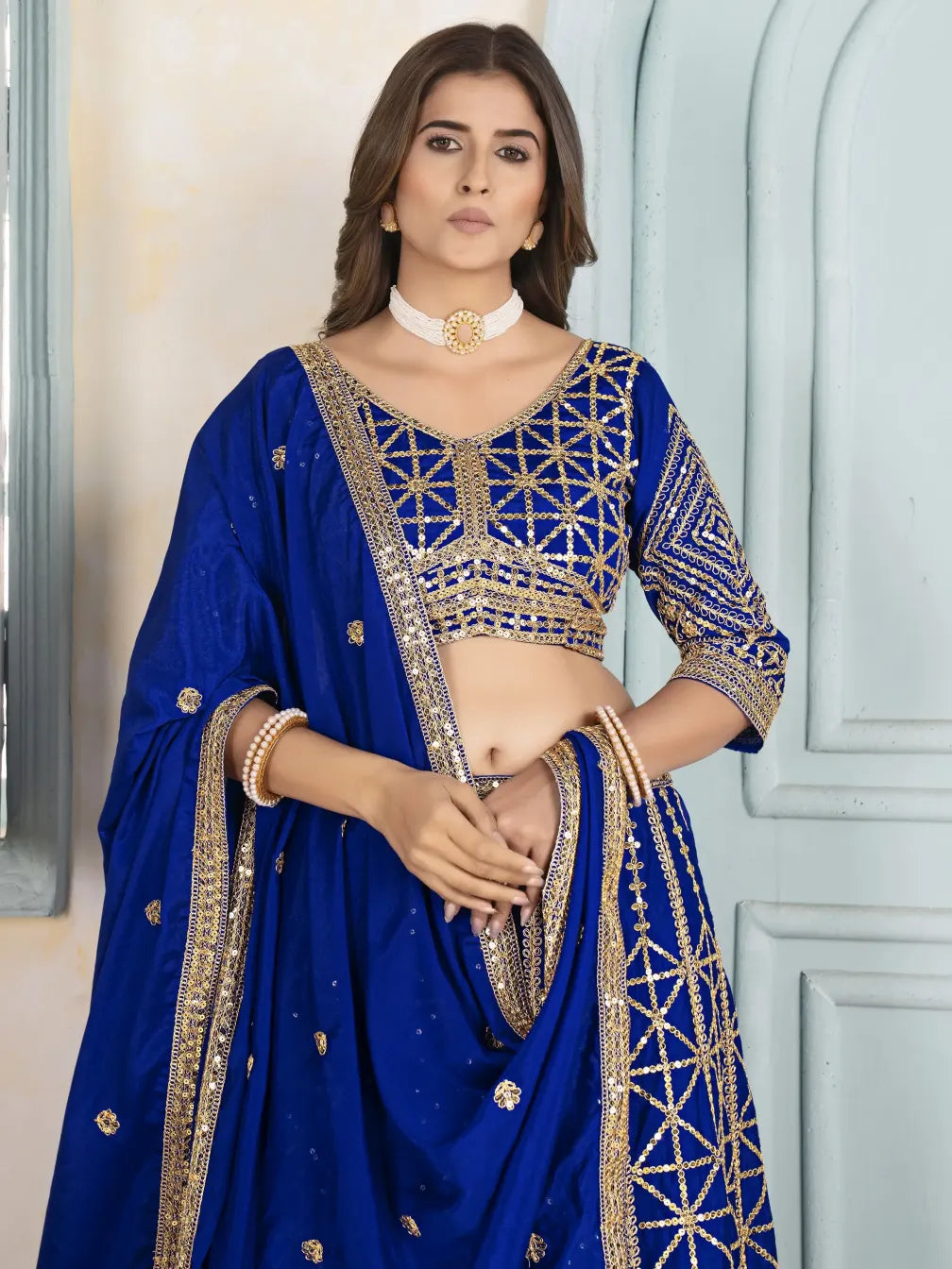Woman wearing a blue embroidered saree with gold details against a light-colored wall.