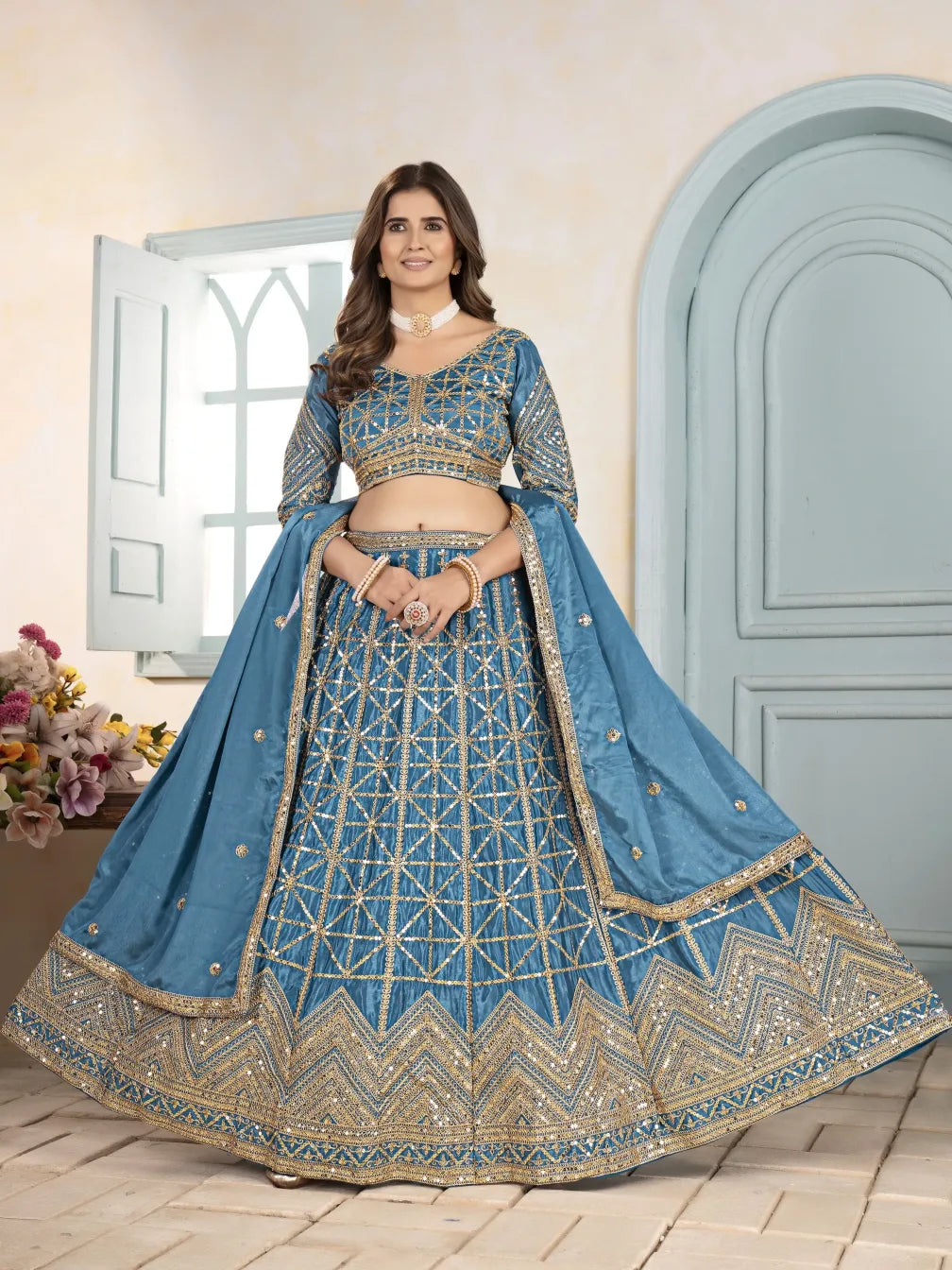 Woman wearing a blue embroidered traditional outfit standing in a room with a light-colored wall and a gray door.