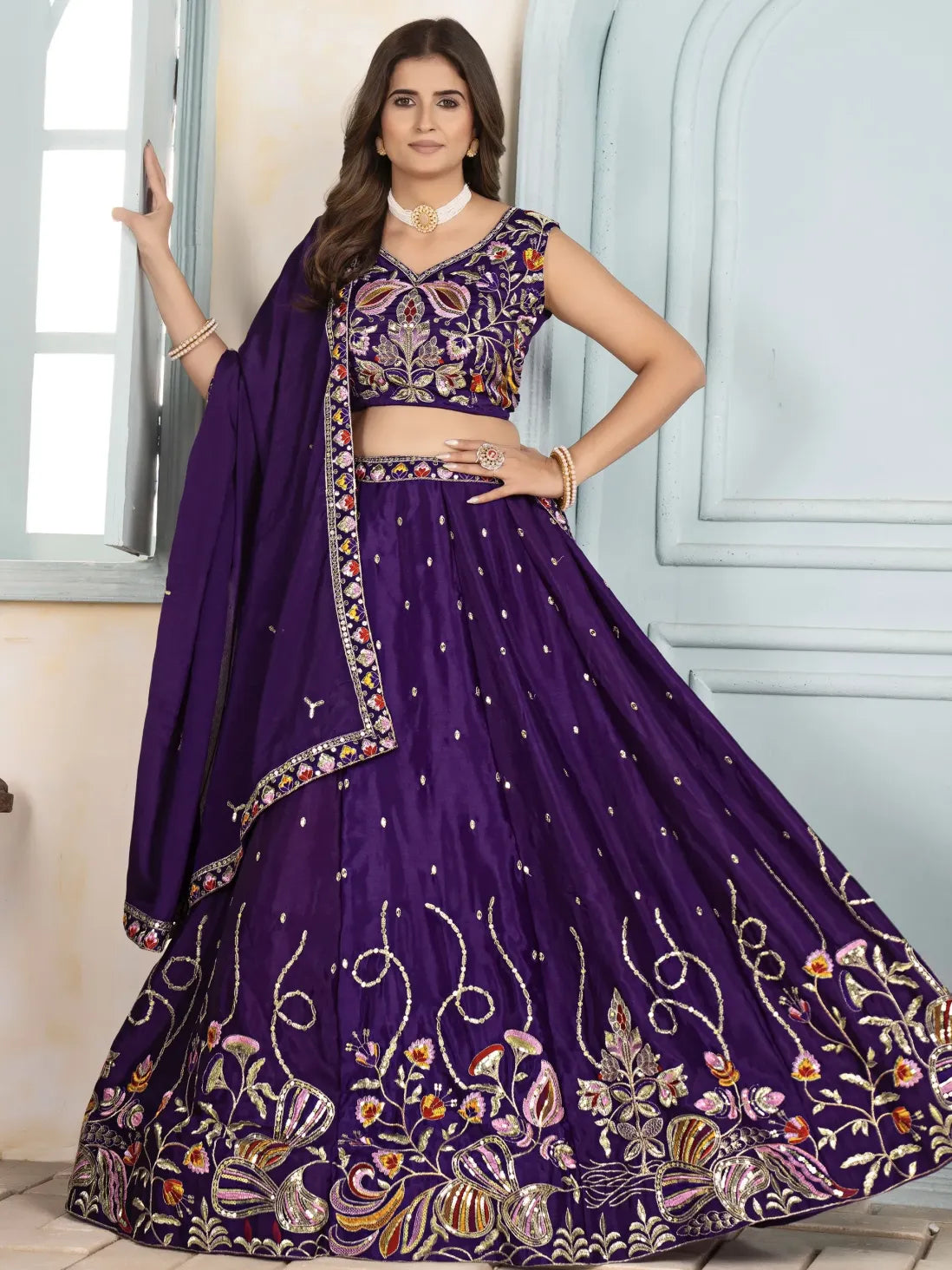 Woman wearing a purple embroidered lehenga with a matching dupatta against a light-colored wall.