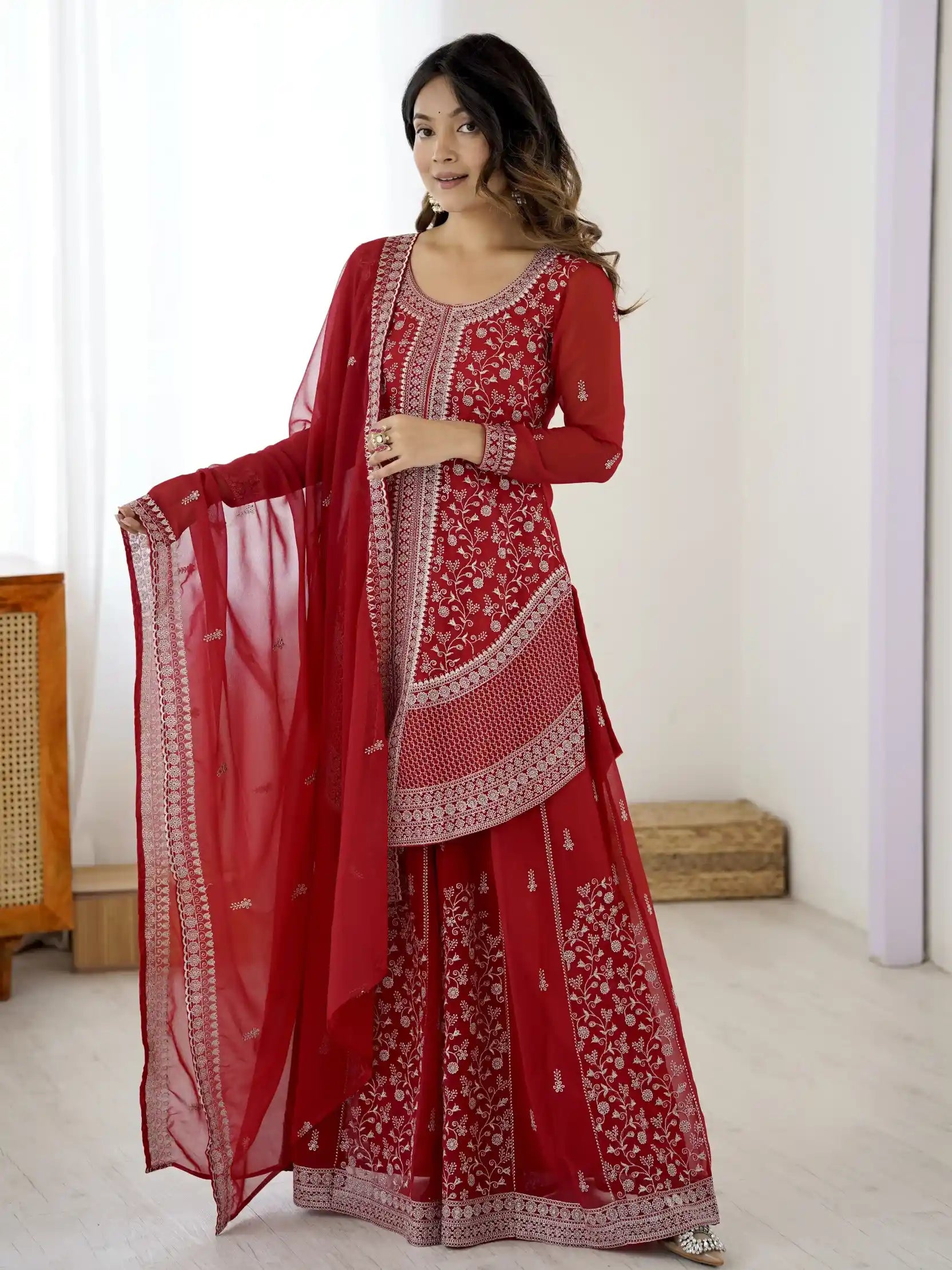 Woman wearing a red embroidered traditional outfit with a dupatta in a minimal indoor setting.