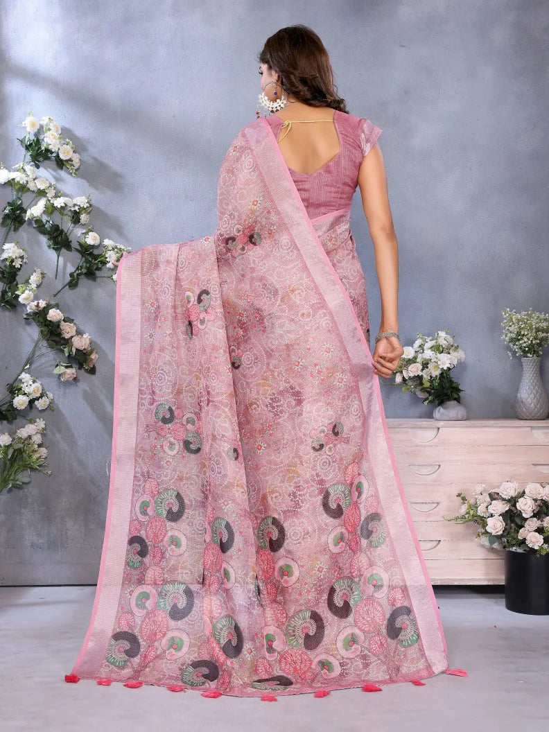 Woman wearing a pink saree with a floral pattern, standing against a gray wall with decorative flowers.
