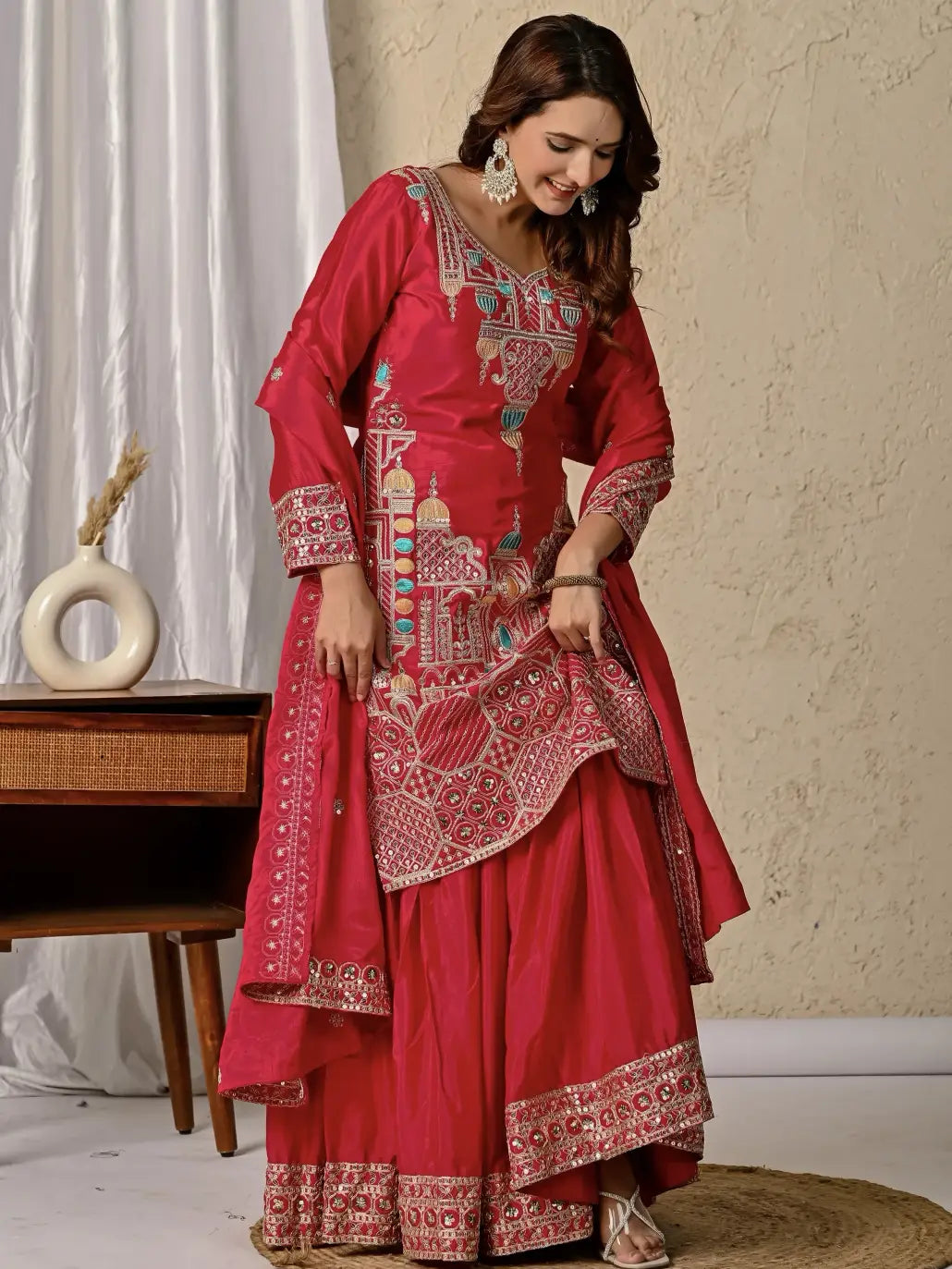 Woman wearing a red traditional outfit with intricate patterns in a room setting.
