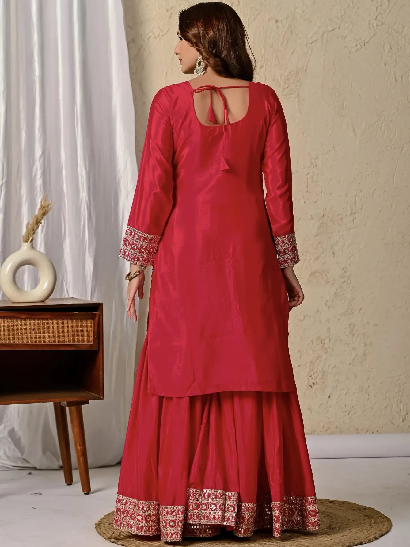 Woman wearing a red traditional outfit with intricate patterns, standing in a room with a decorative cabinet and white curtain.