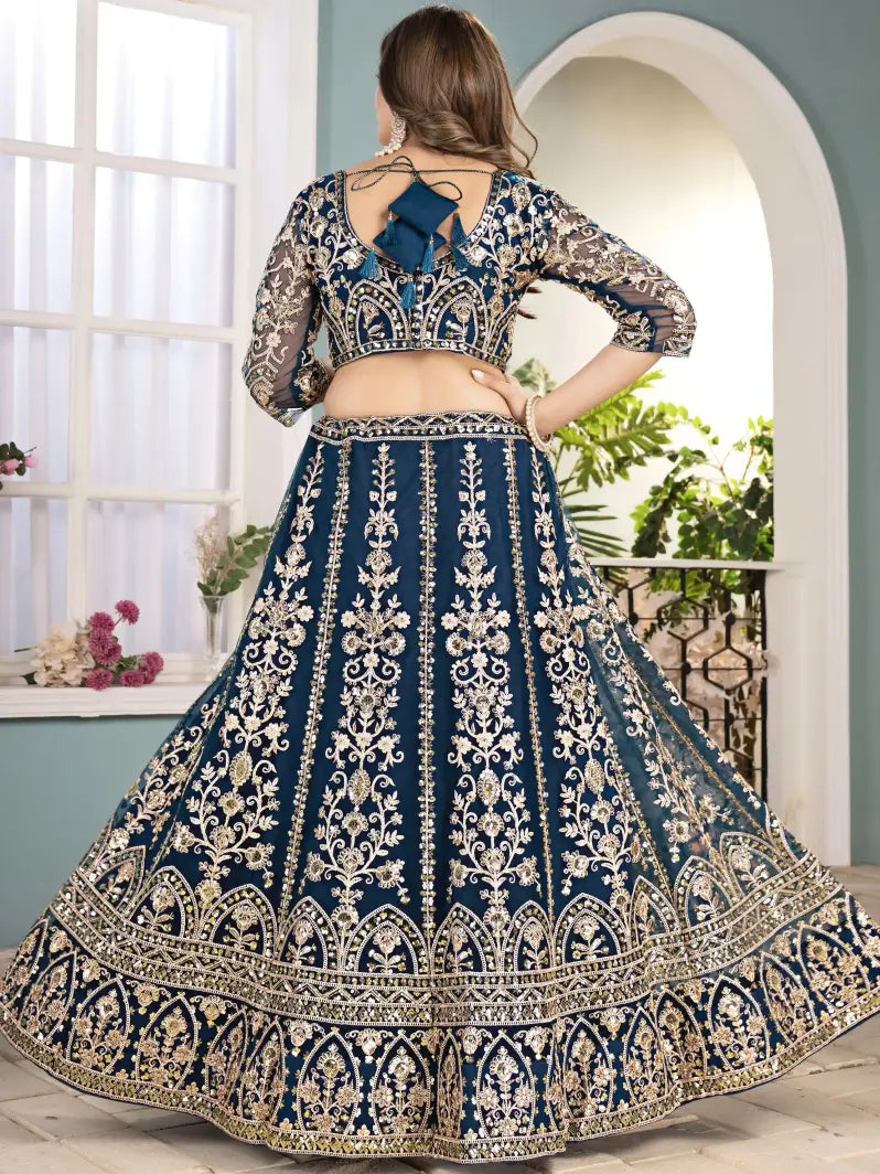 Woman wearing a blue and gold embroidered traditional outfit in a room with a window and plants.