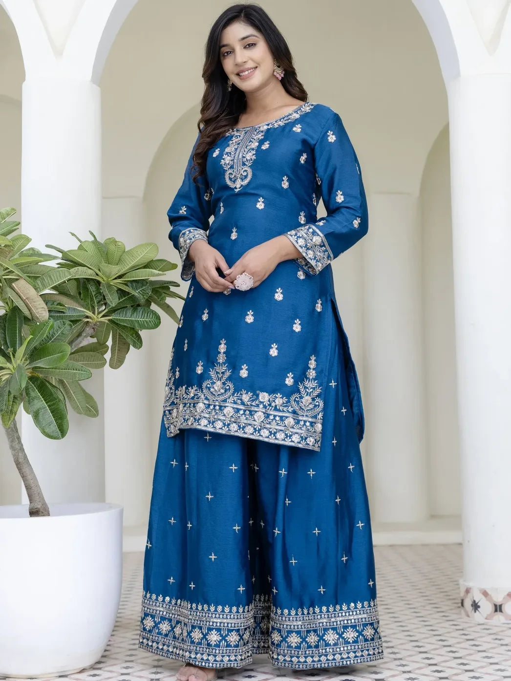 Woman wearing a blue embroidered traditional outfit standing in a decorative setting with plants.