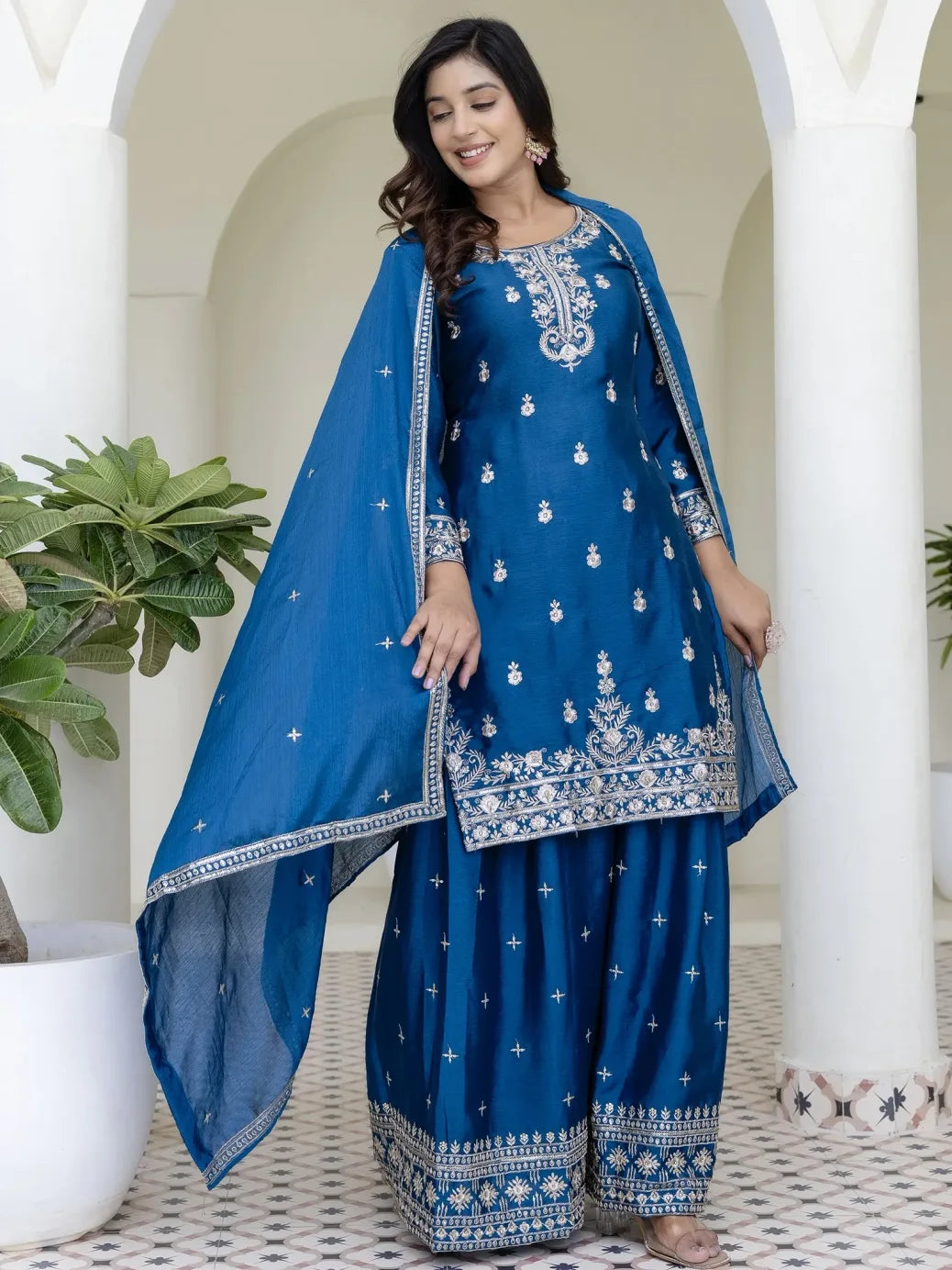 Woman wearing a blue embroidered traditional outfit with a matching dupatta indoors.