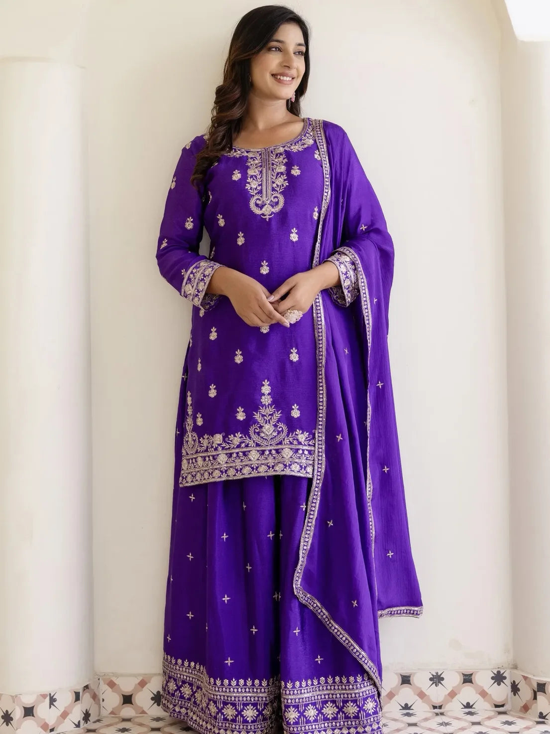 Woman wearing a purple traditional outfit with white embroidery against a white curtain background