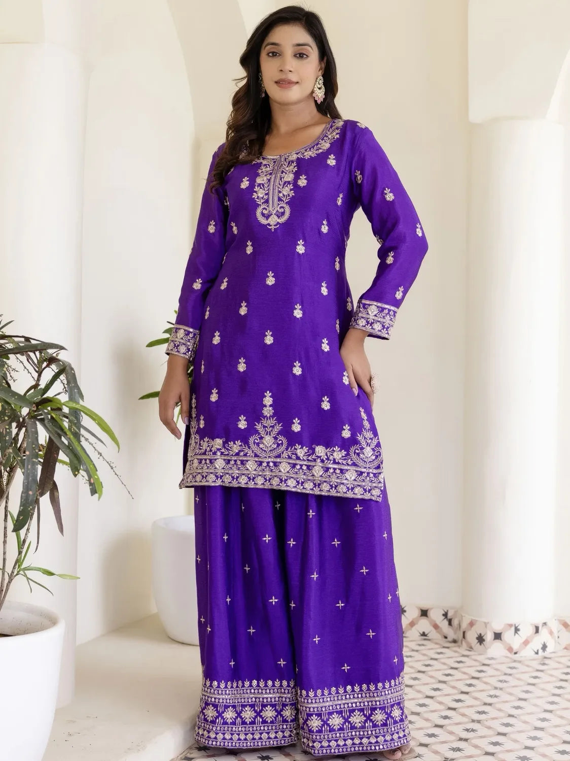 Woman wearing a purple embroidered traditional outfit in a minimalistic room.