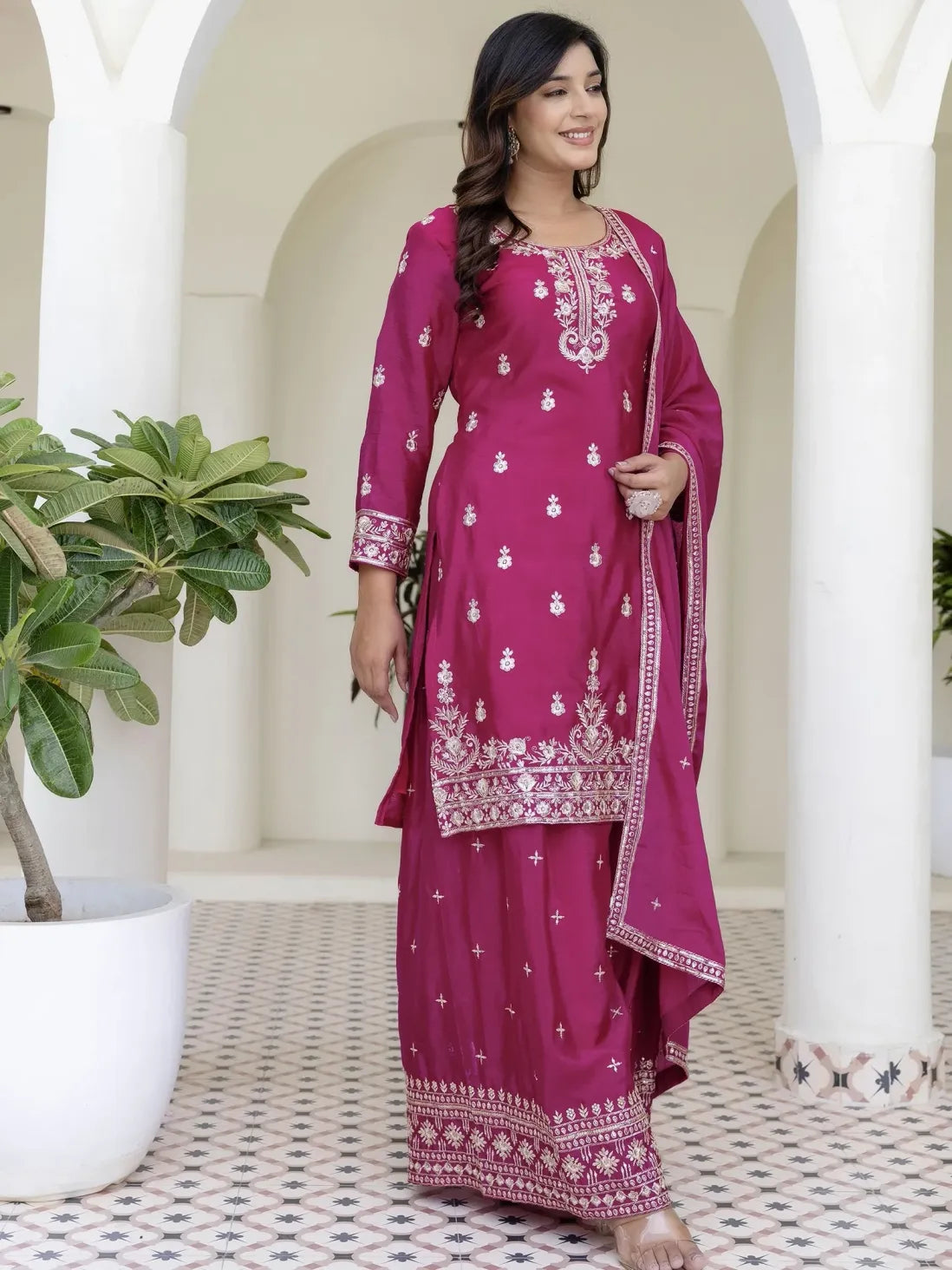 Woman in a pink traditional outfit with white embroidery standing in a decorative interior setting.