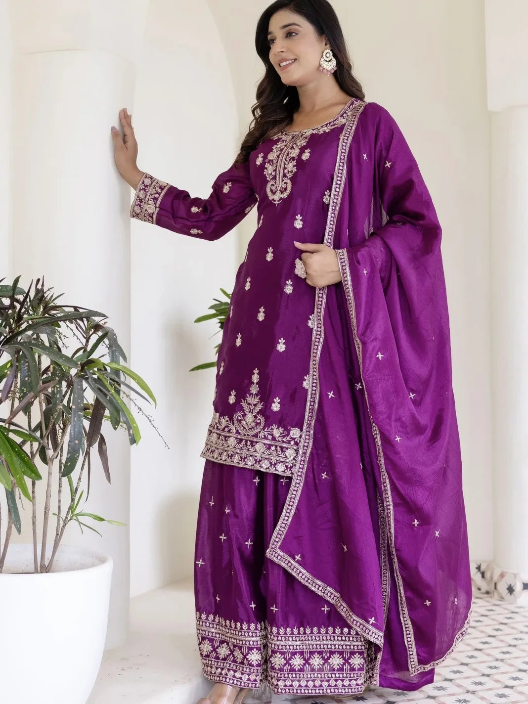 Woman wearing a purple traditional outfit with white embroidery in a minimal indoor setting.