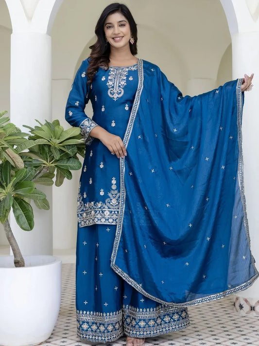 Woman wearing a blue embroidered traditional outfit with a matching dupatta, standing indoors.
