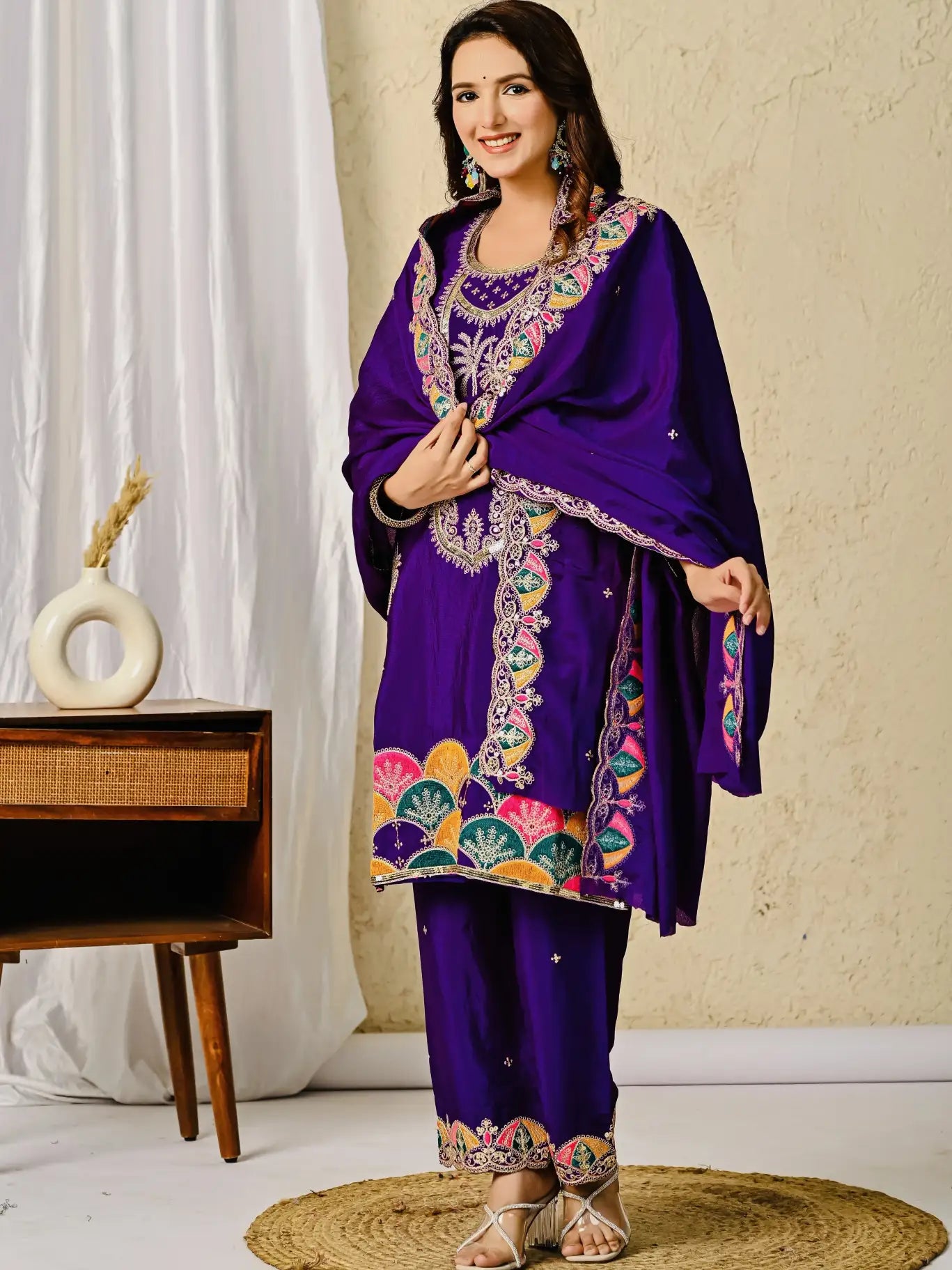 Woman wearing a purple embroidered traditional outfit in a room with a white curtain and wooden table.