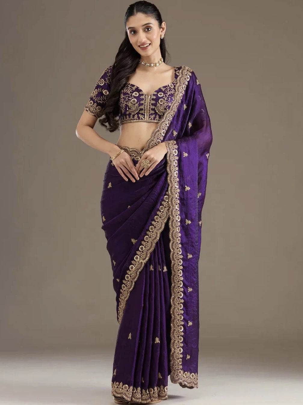 Woman wearing a purple saree with gold embroidery on a plain background