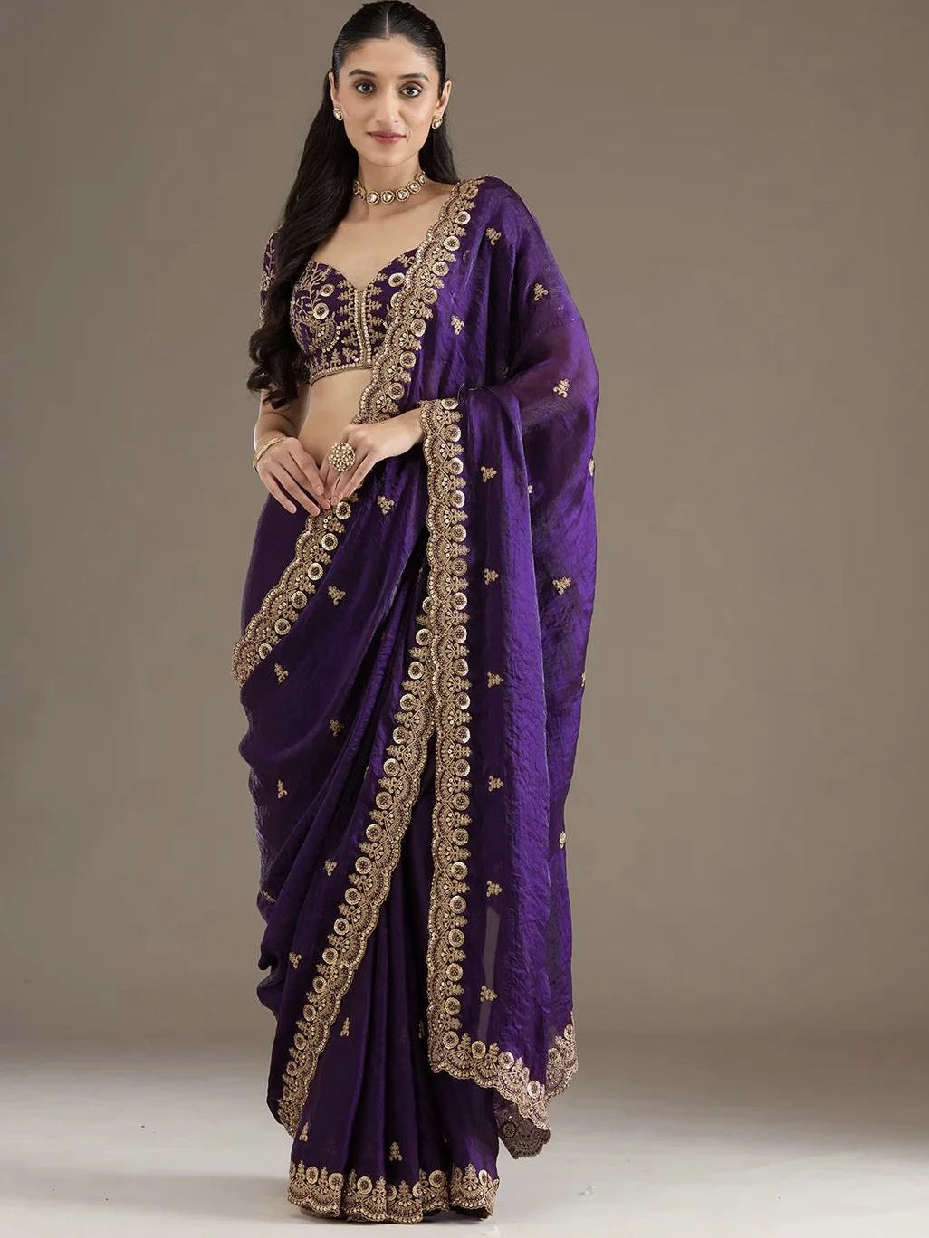 Woman wearing a purple saree with gold embroidery on a plain background