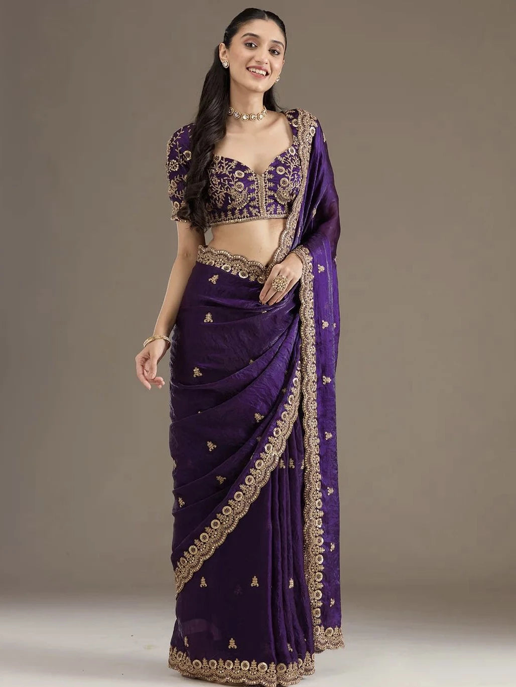 Woman wearing a purple saree with gold embroidery on a plain background