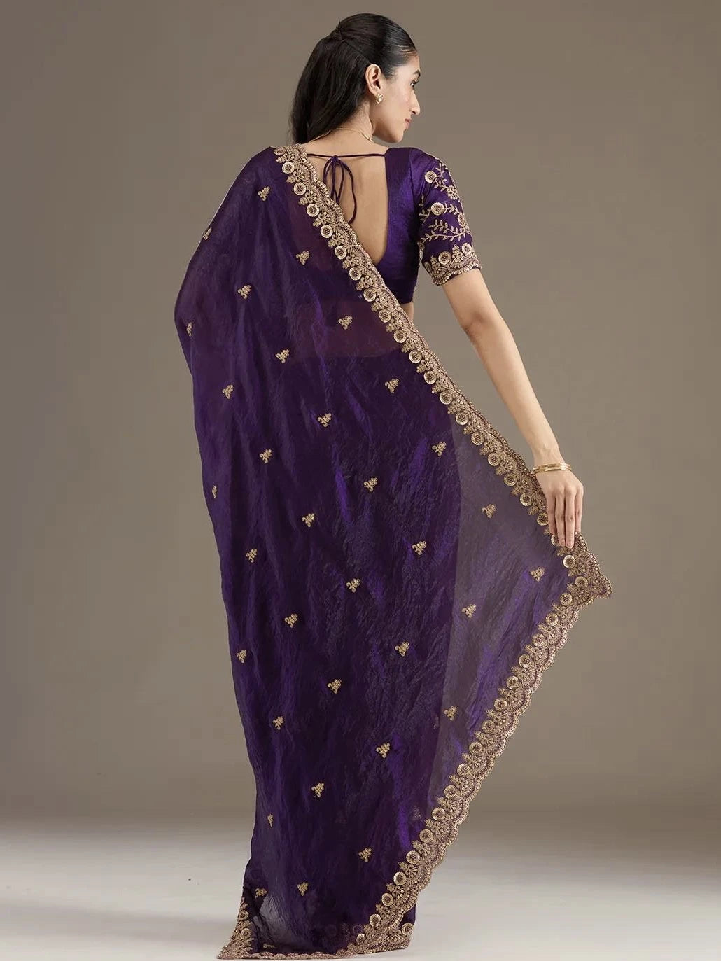 Woman wearing a purple saree with gold embroidery on a plain background