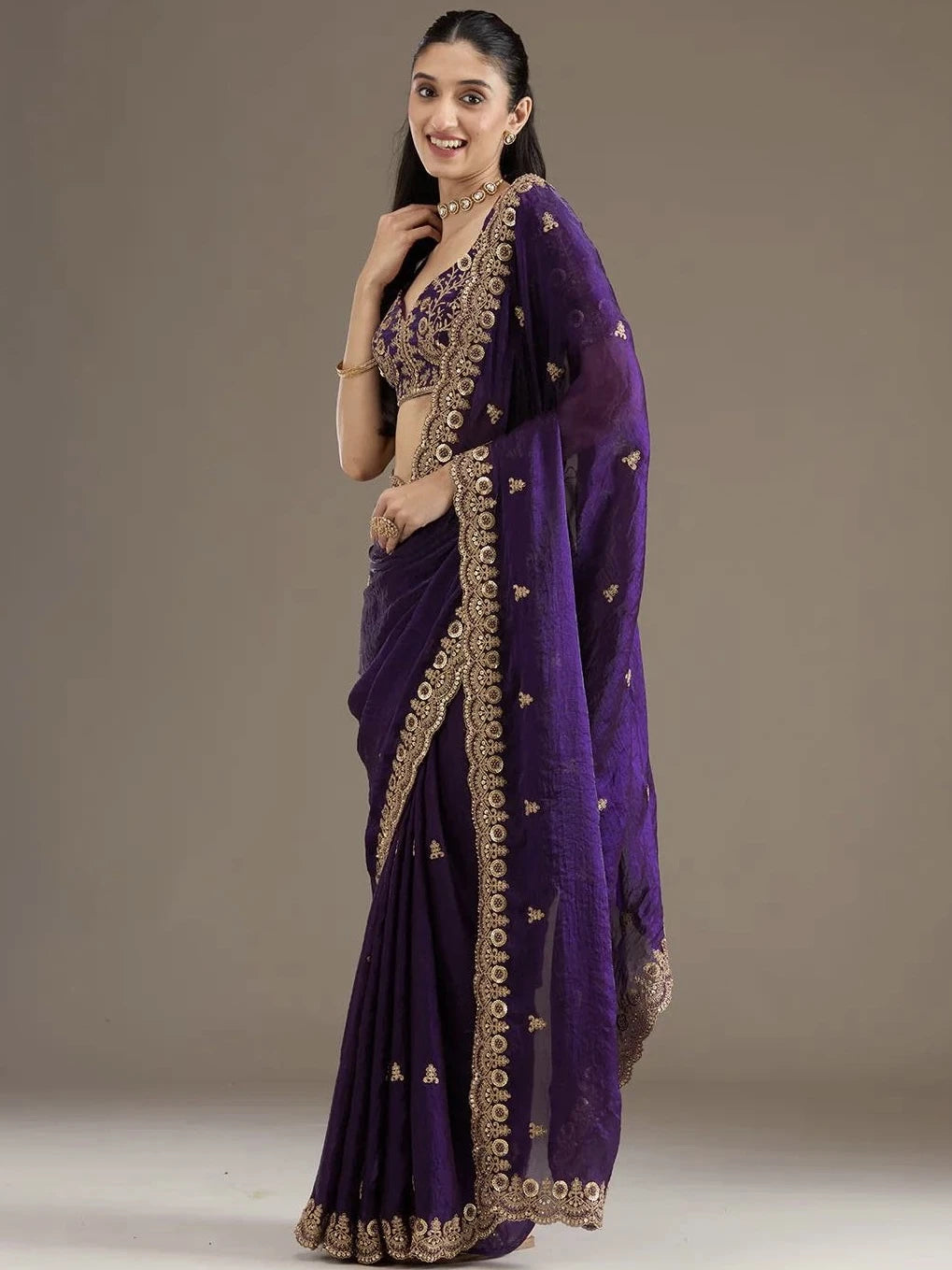 Woman wearing a purple saree with gold embroidery on a plain background