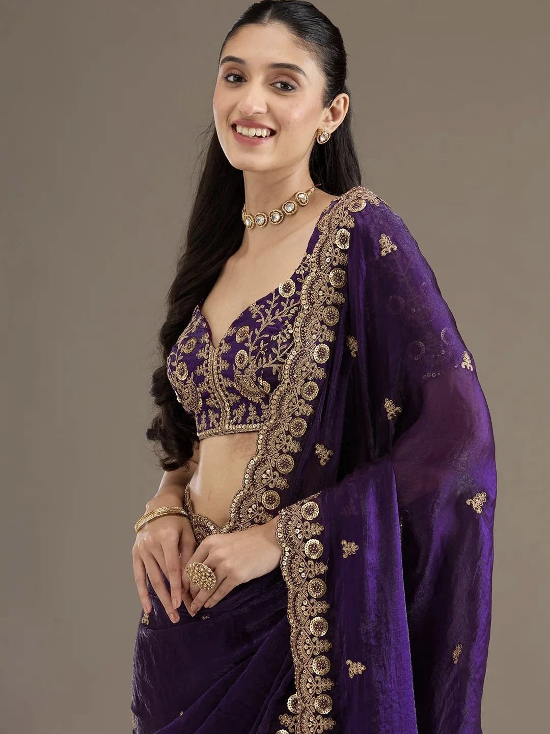 Woman wearing a purple saree with gold embroidery against a brown background