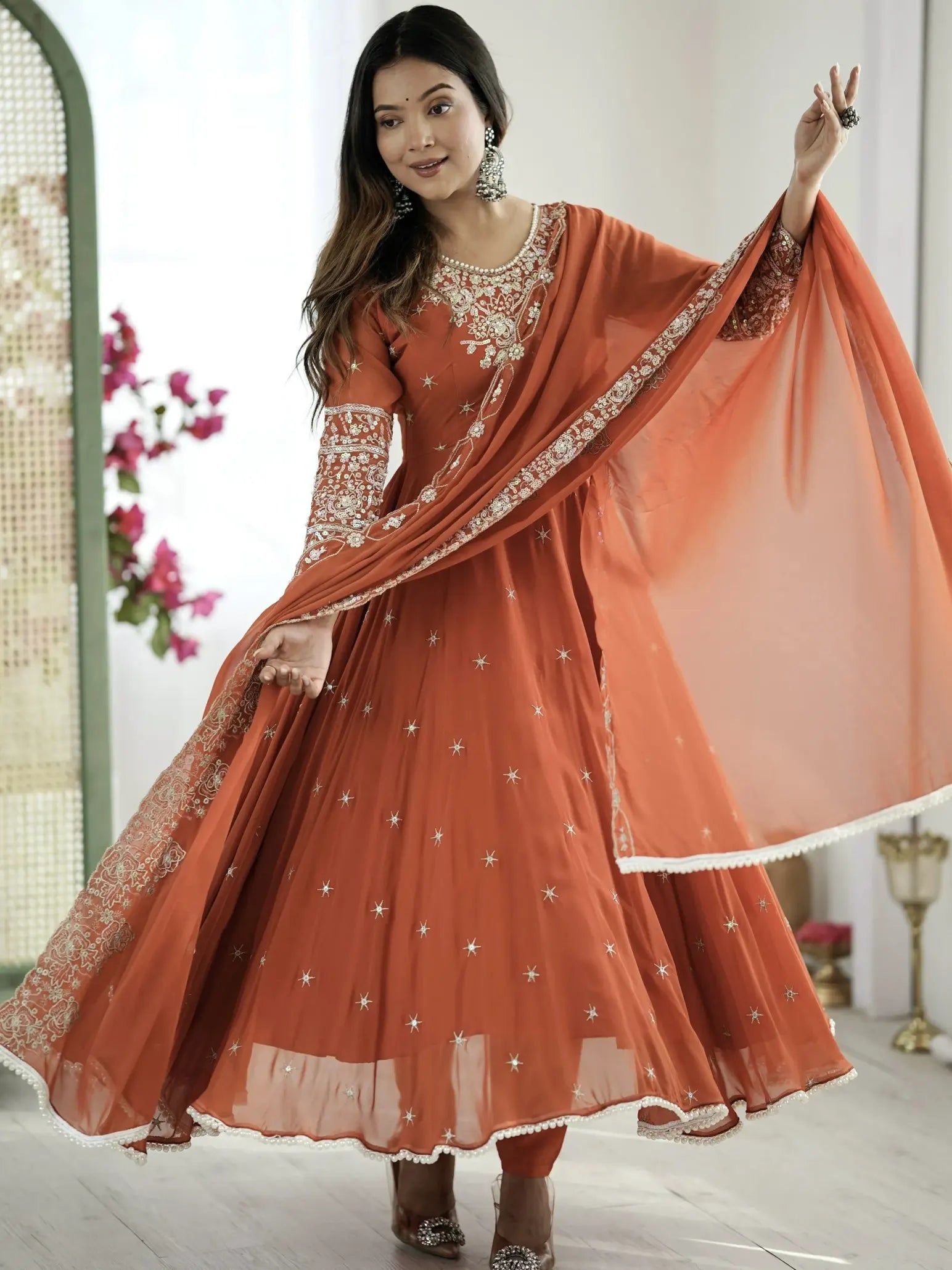 Woman in an orange traditional outfit with a dupatta, standing indoors.