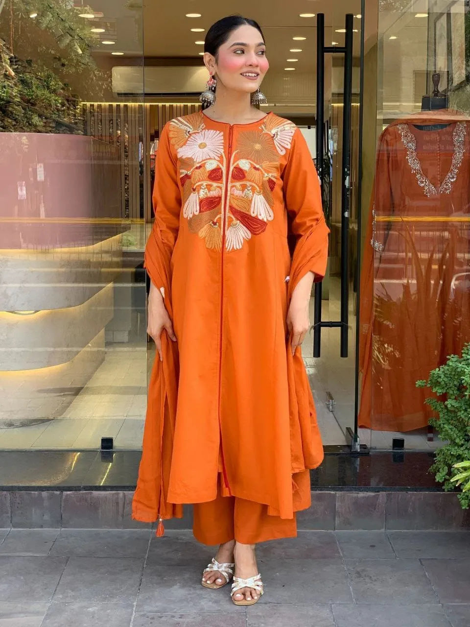 Woman wearing an orange traditional outfit with intricate designs in front of a store.