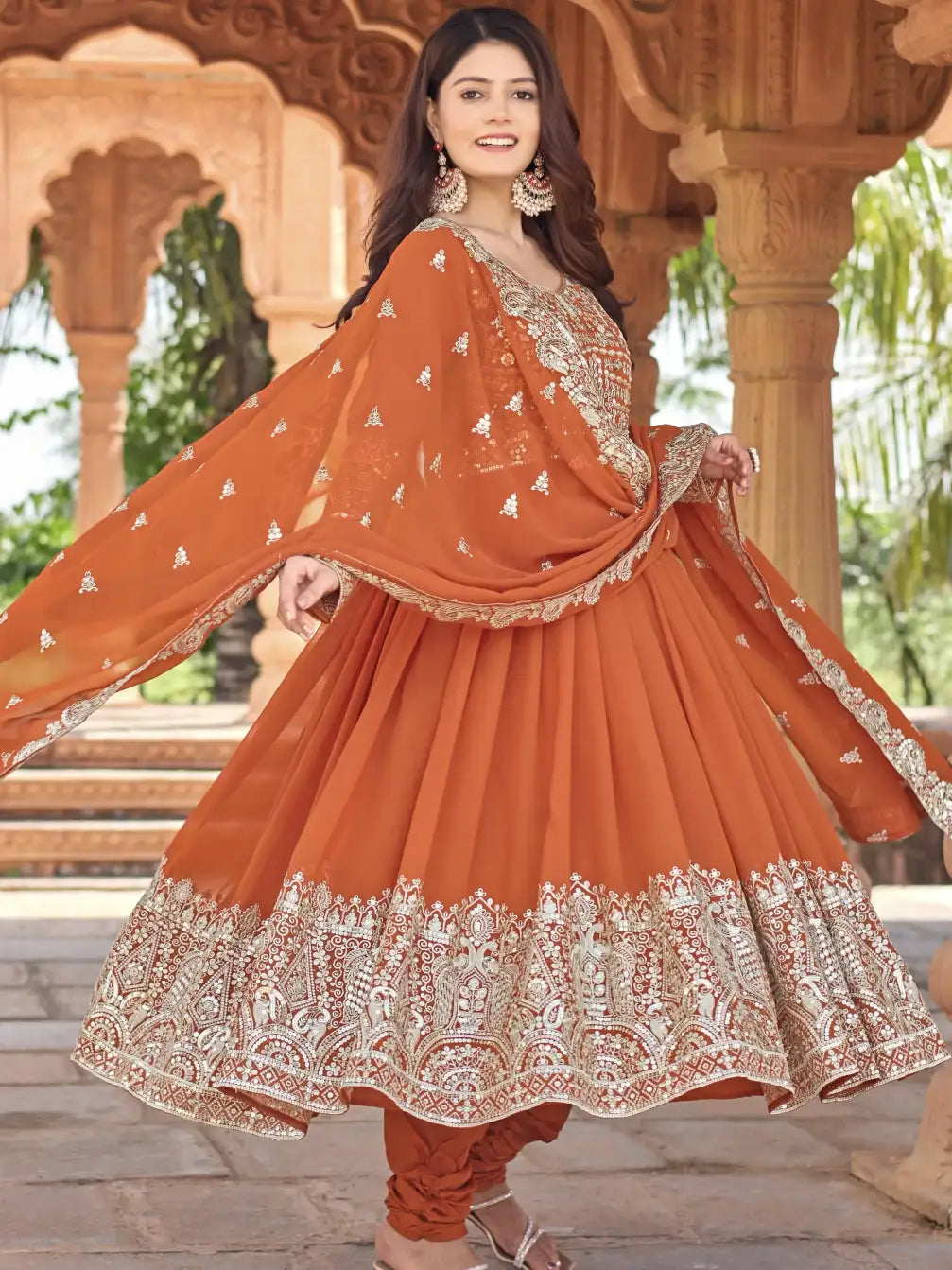 Woman wearing an orange traditional outfit with intricate embroidery in a decorative setting.