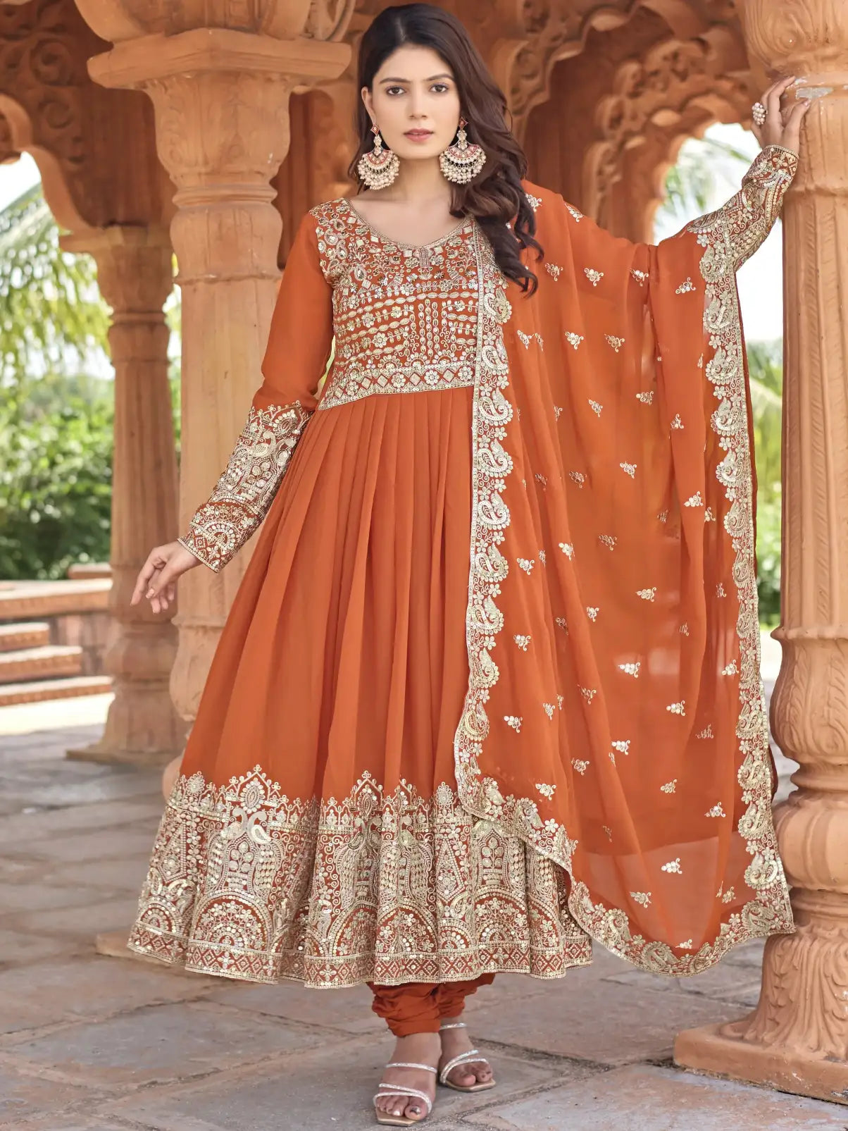 Woman wearing an orange traditional outfit with intricate embroidery in a decorative setting.