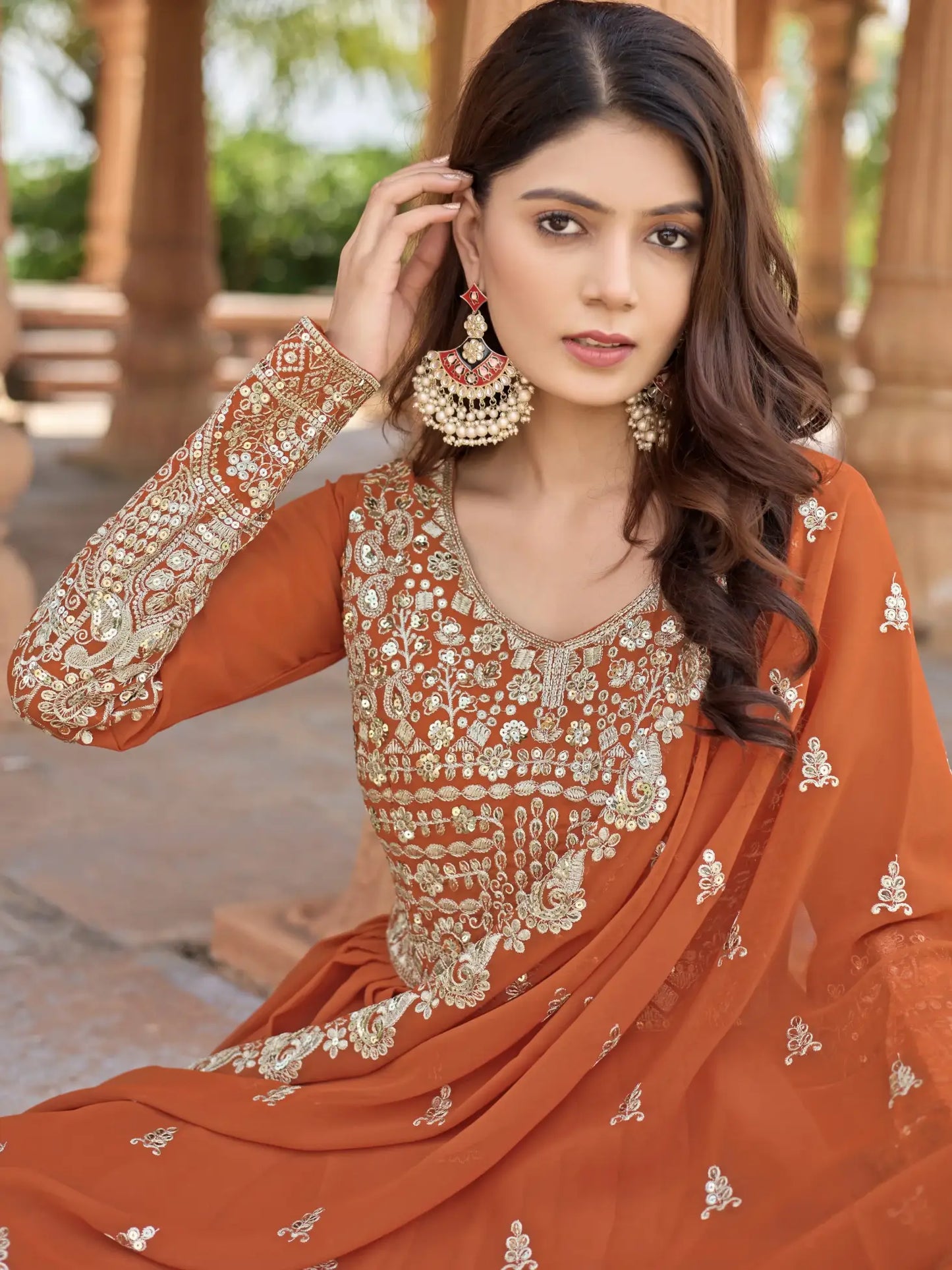 Woman wearing an orange embroidered traditional outfit with gold jewelry outdoors.