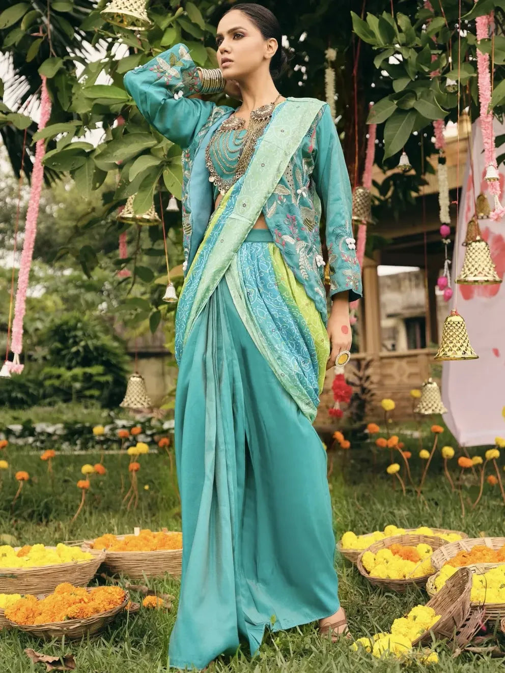Woman in a traditional turquoise saree with floral decorations and hanging bells in the background