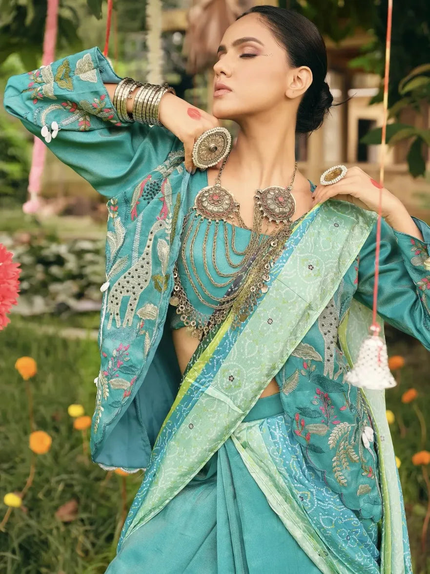 Woman in a traditional turquoise saree with floral patterns and jewelry, standing outdoors.