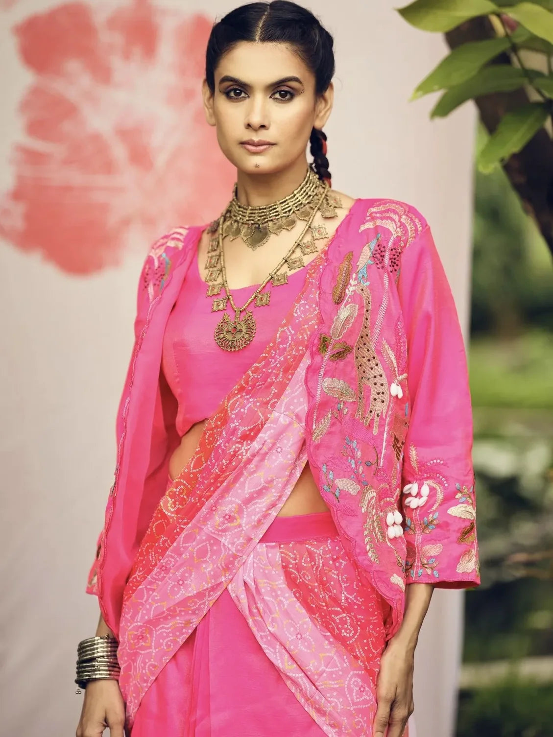 Woman wearing a pink saree with floral patterns and gold jewelry against a neutral background.