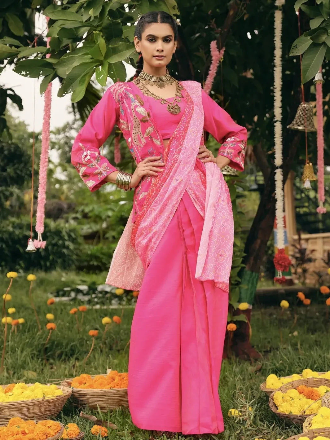 Woman in a pink saree standing outdoors with greenery and flowers in the background