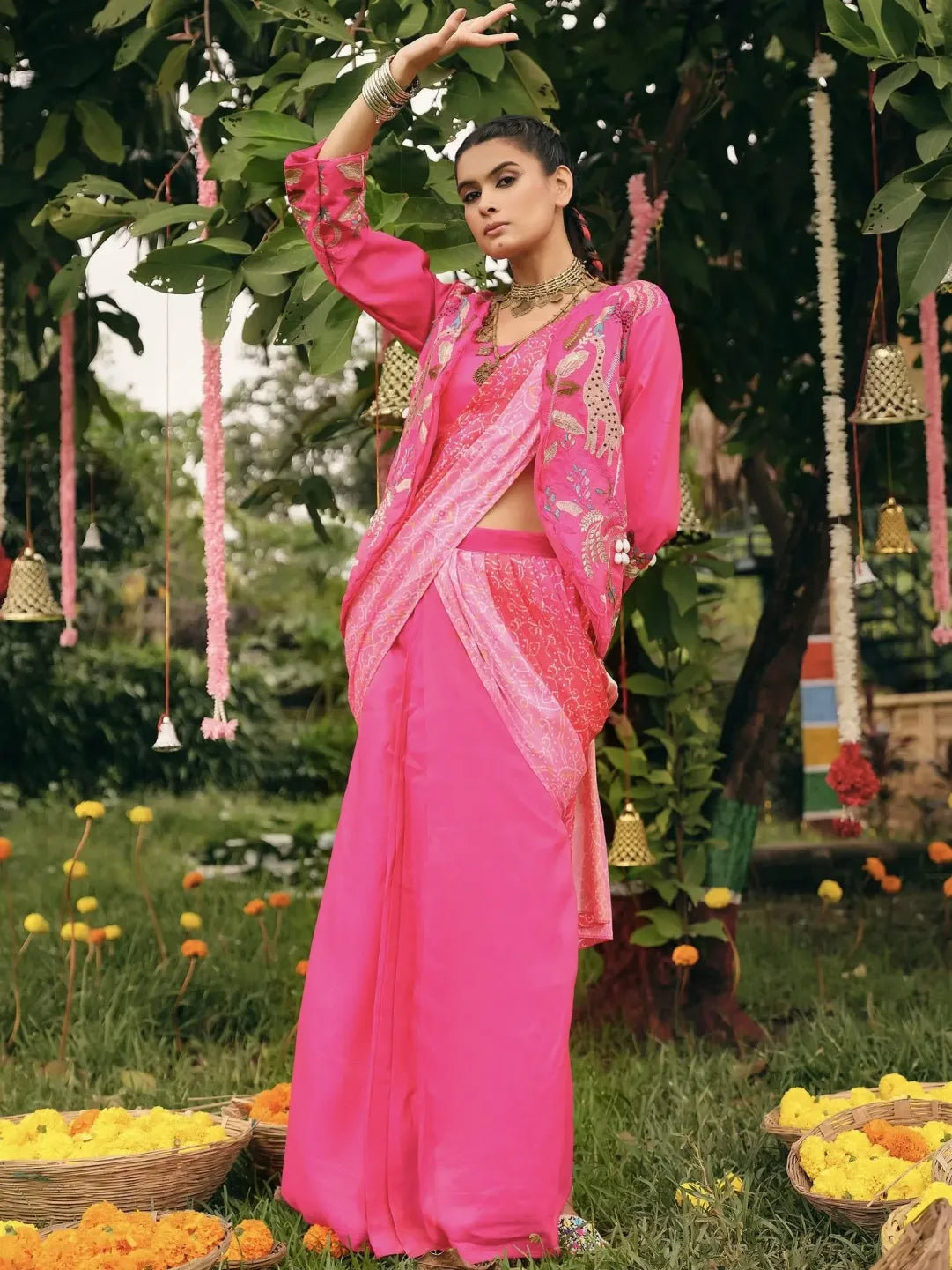 Woman in a bright pink saree standing outdoors with greenery and decorations around.