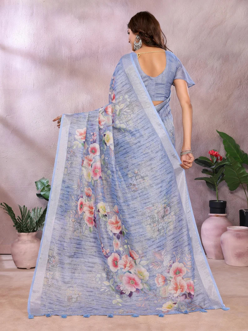 Woman wearing a light blue saree with floral patterns in a room with plants.