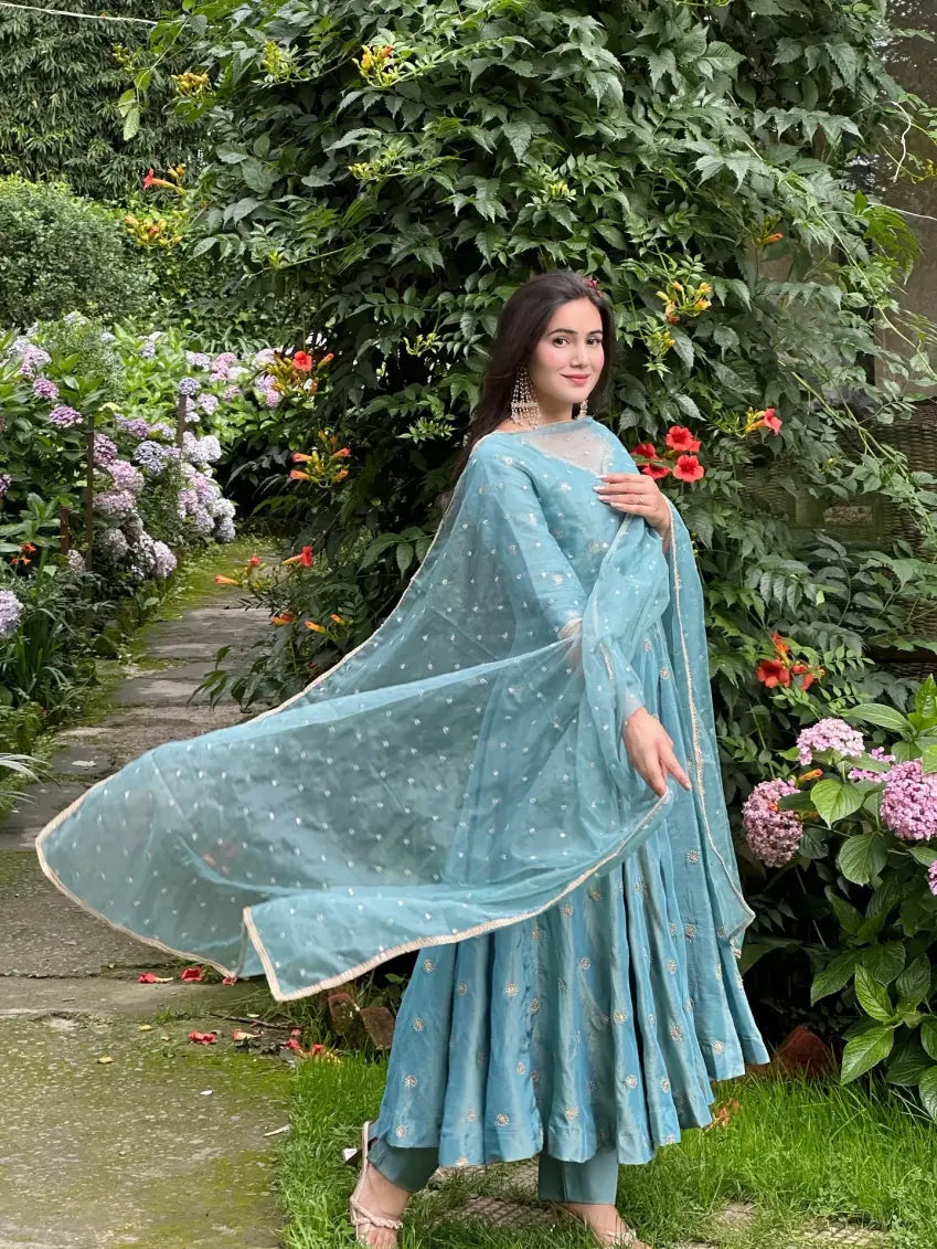 Woman in a light blue traditional outfit standing in a garden with flowers and greenery.