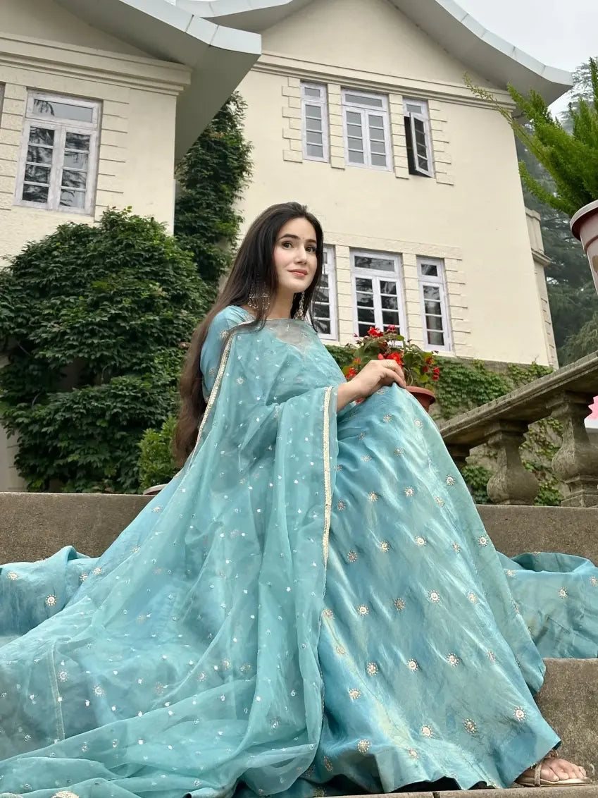 Woman in a light blue saree standing outdoors with a building in the background